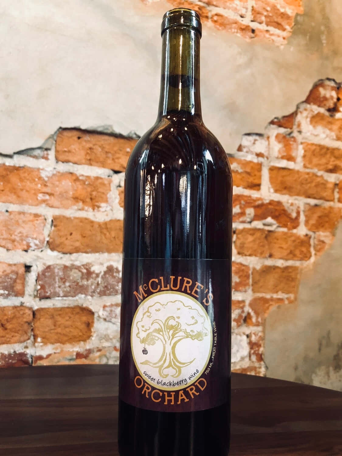 Blackberry Wine 桌布 壁紙 — 一個深色酒瓶帶有樹木標誌，放在木桌上，背景是質樸的磚牆。
