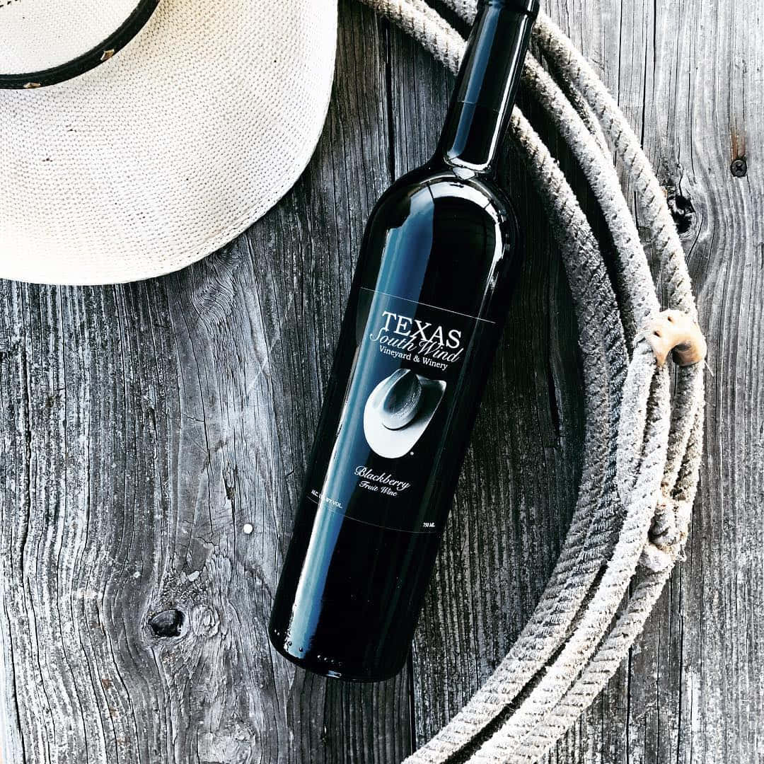 Blackberry Wine 桌布 壁紙 — 一個深色酒瓶標有Texas South Wind，放在風化木頭上，旁邊有一頂牛仔帽和一條捲起的繩索。