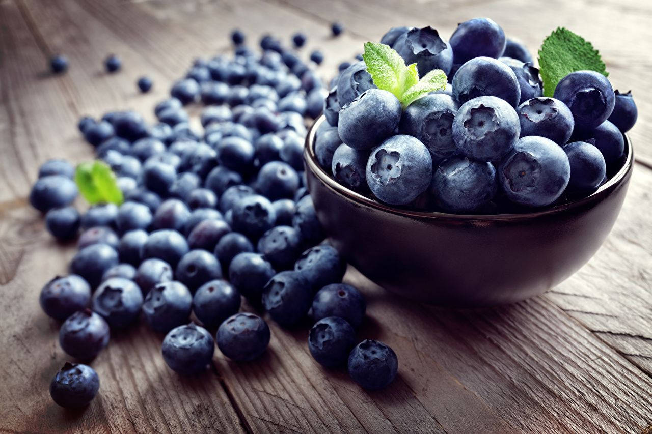 Fruits 桌布 壁紙 — 一個深色陶瓷碗裝滿了飽滿的藍莓和新鮮薄荷葉，放在一個質樸的木桌上，周圍散落著一些莓果。