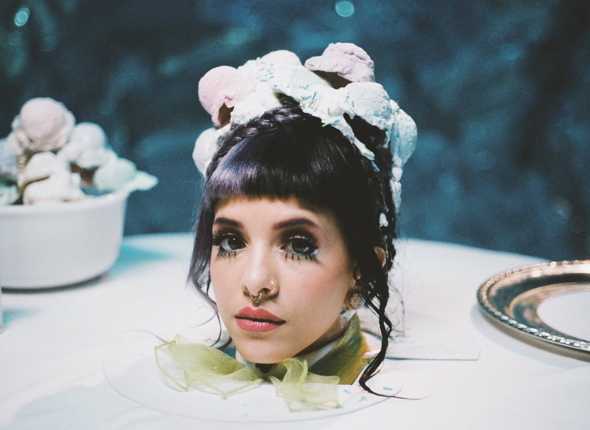Melanie 桌布 壁紙 — 一個深色頭髮、化著大膽妝容的人戴著一個奇特的冰淇淋筒頭飾，坐在一張放有一碗冰淇淋球和一個銀盤的桌子旁。