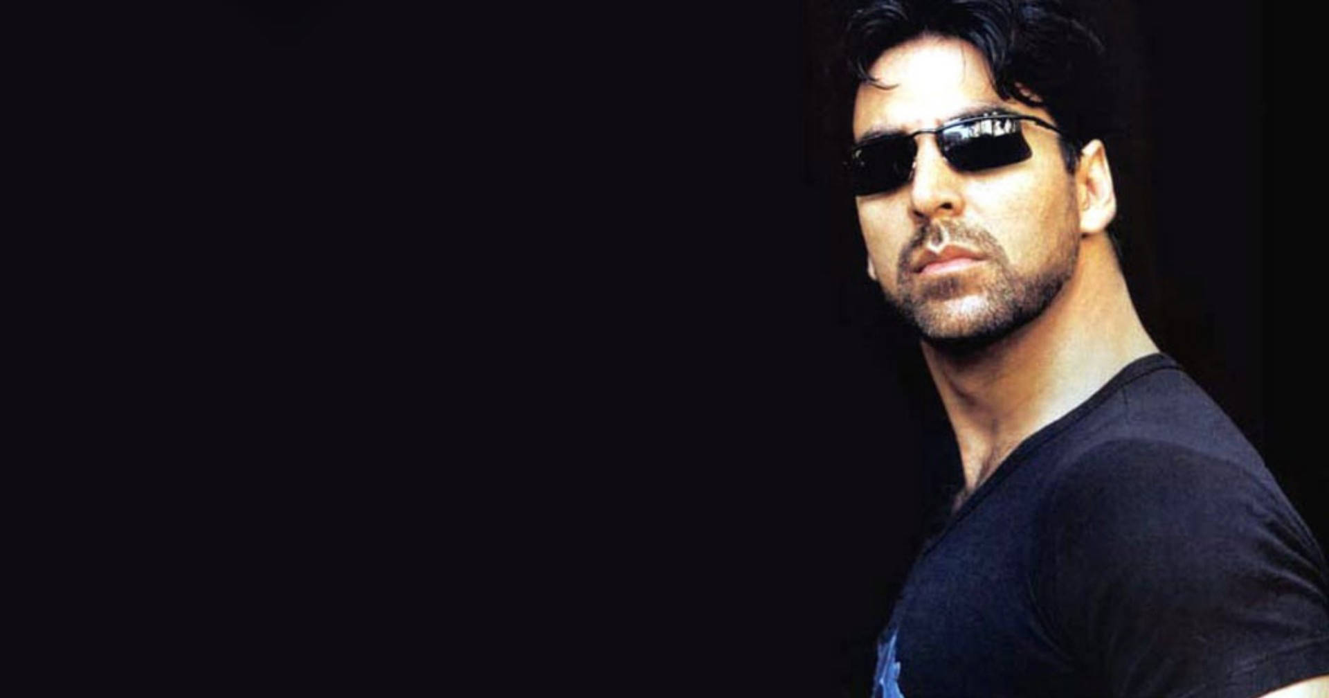 Akshay Kumar 桌布 壁紙 — 一個深色頭髮、戴太陽眼鏡的男人，穿著黑色T恤，在黑色背景前凝視著。