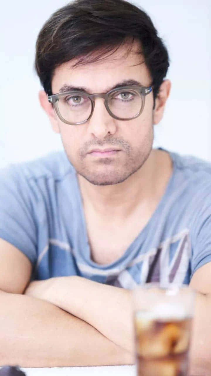Aamir Khan 桌布 壁紙 — 一個深色頭髮、戴眼鏡的男人，雙手擱在桌上，旁邊有杯飲品，專注地凝視鏡頭。