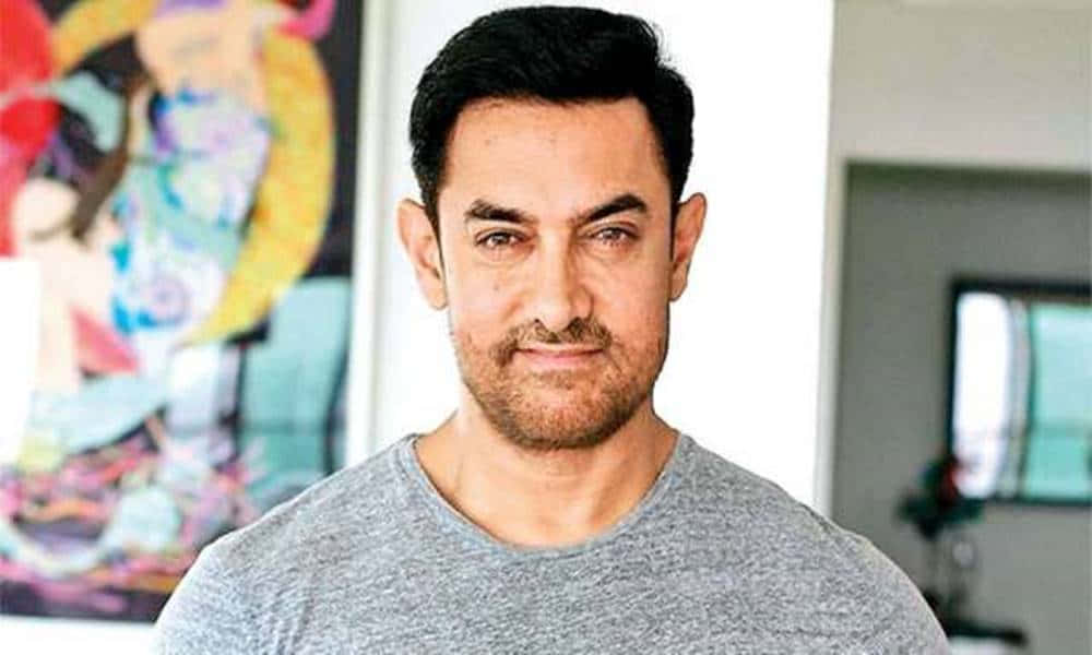 Aamir Khan 桌布 壁紙 — 一個深色頭髮、淺色鬍鬚的男人，穿著灰色T恤，站在室內一幅色彩繽紛的抽象畫附近。