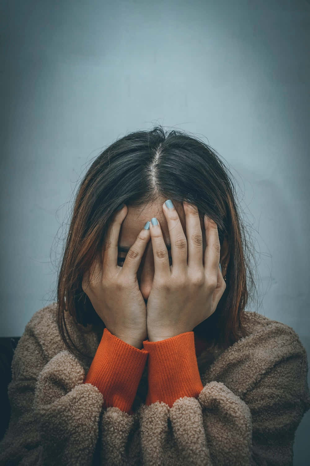 Sad Profile 桌布 壁紙 — 一個深色頭髮、淺藍色指甲的人用雙手遮住臉，穿著一件帶橙色袖口的棕色抓絨外套，背景是柔和的灰色。