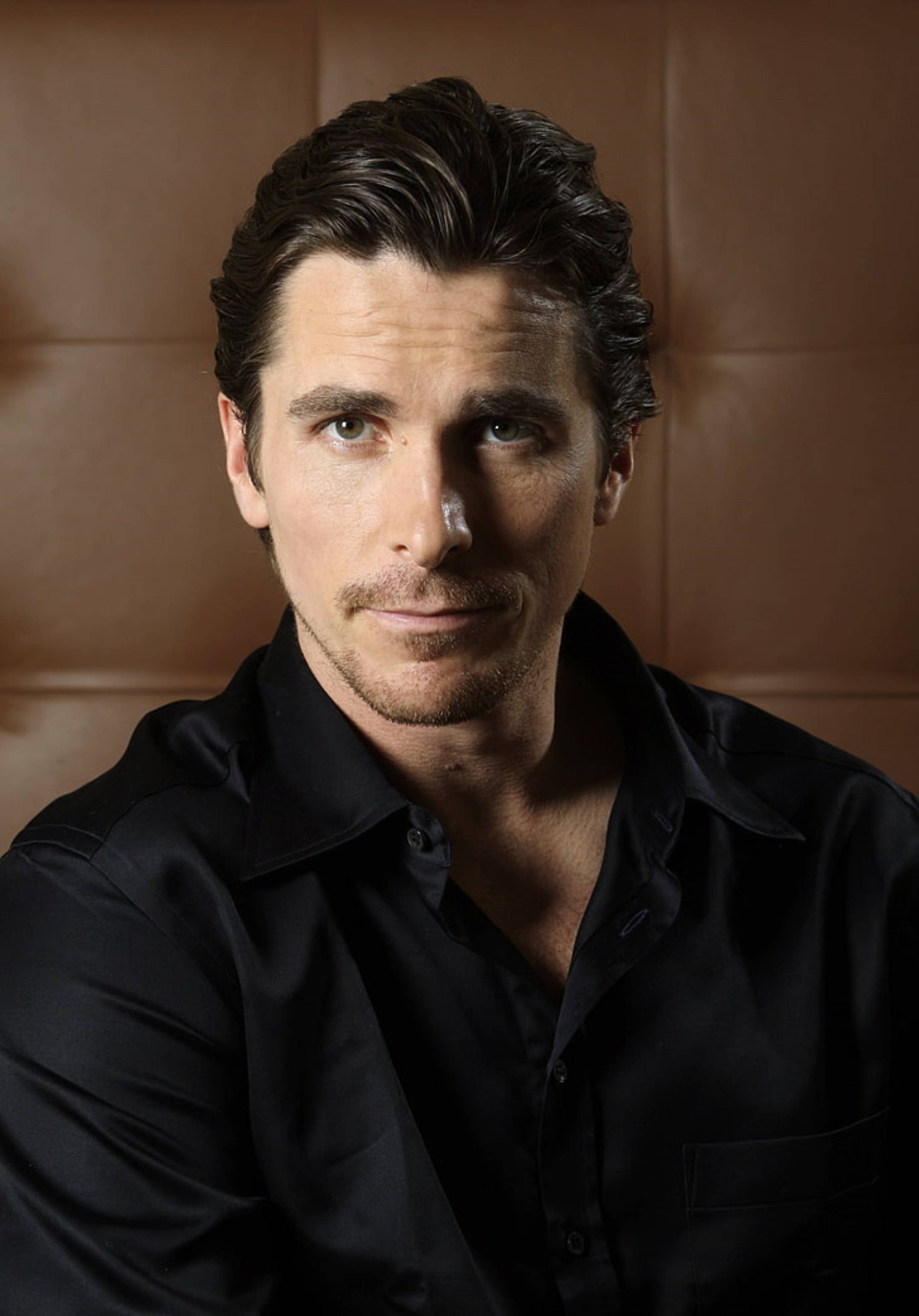 Christian Bale 桌布 壁紙 — 一個深色頭髮、留著輕微鬍鬚的男人，穿著黑色襯衫，背景是棕色皮革。