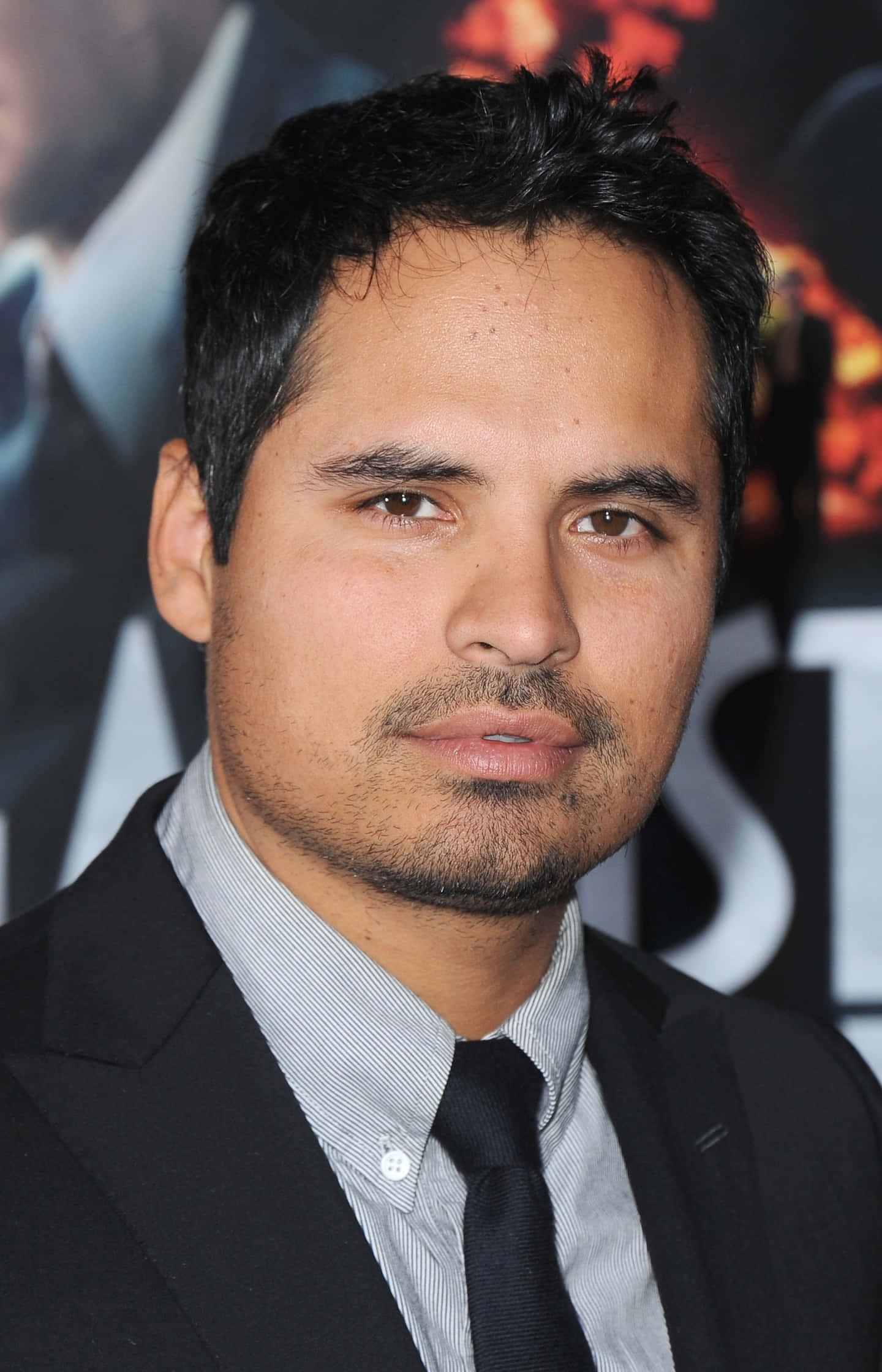 Michael Peña 桌布 壁紙 — 一個深色頭髮、留鬍渣的男人，身穿黑色西裝打領帶，背景是模糊的電影海報，近距離肖像。