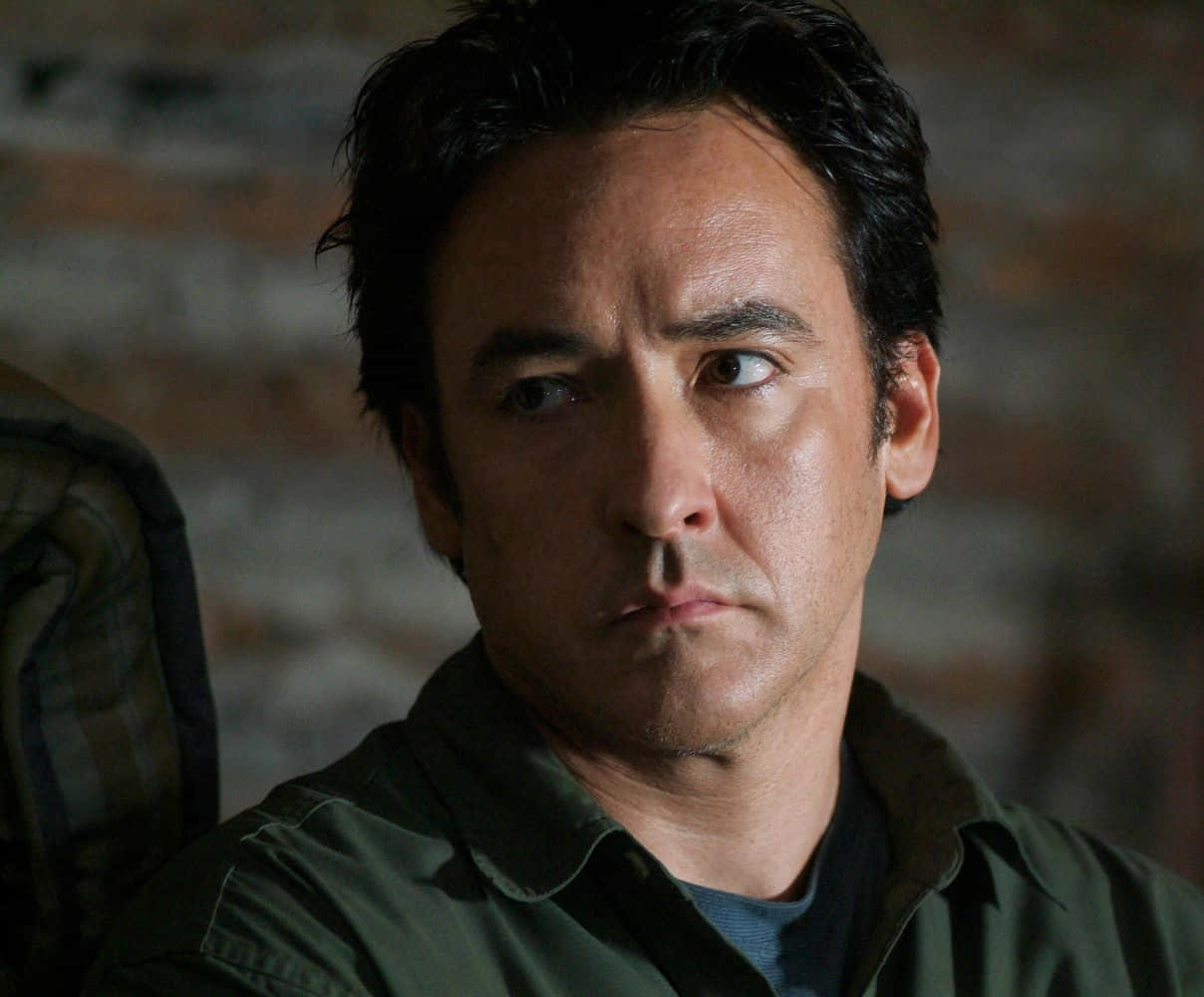 John Cusack 桌布 壁紙 — 一個深色頭髮、眼神銳利的男人，在模糊的磚牆背景下向前凝視。