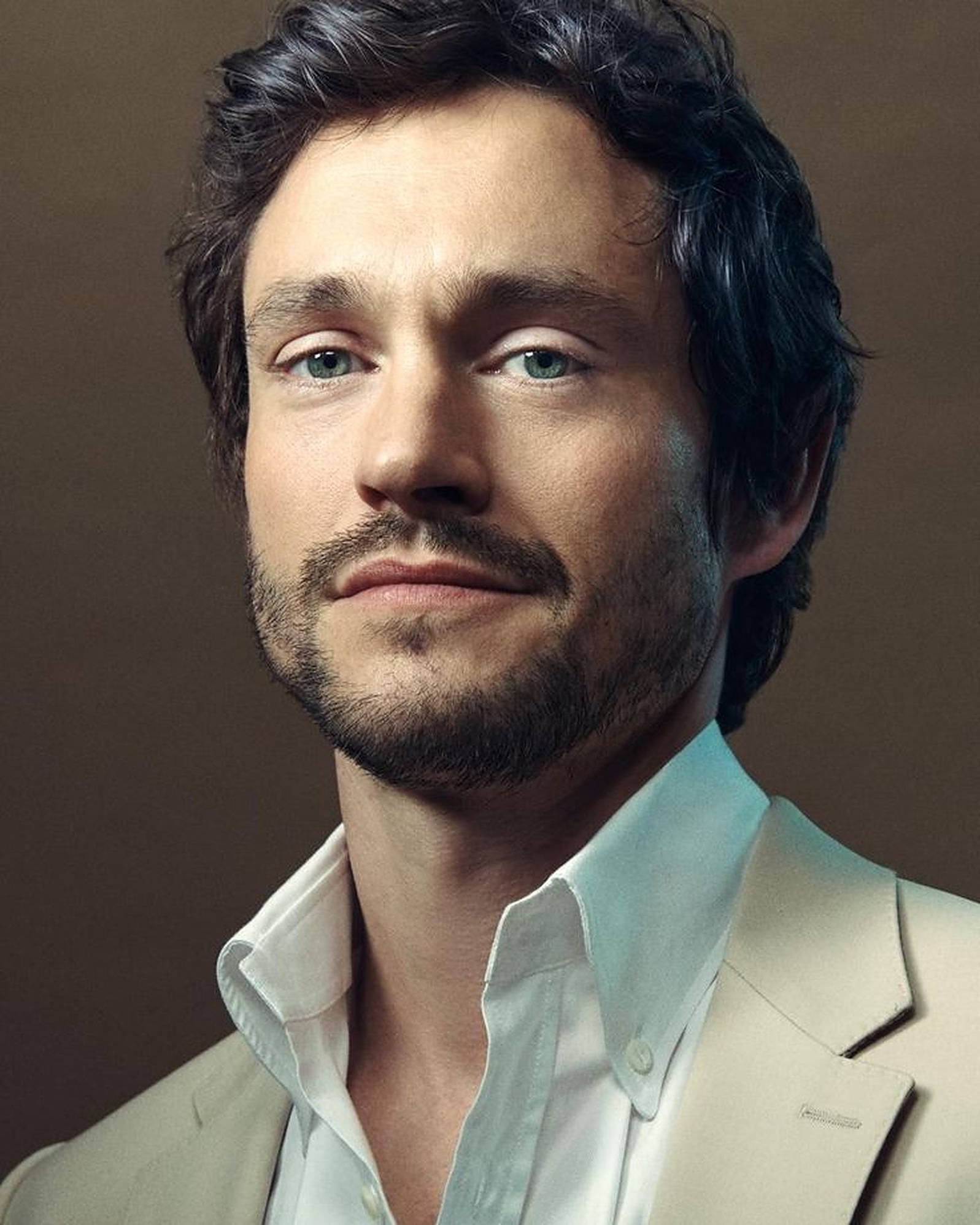 Hugh Dancy 桌布 壁紙 — 一個深色頭髮、藍色眼睛的男人的特寫肖像，他穿著淺米色西裝和白色襯衫，背景是棕色。