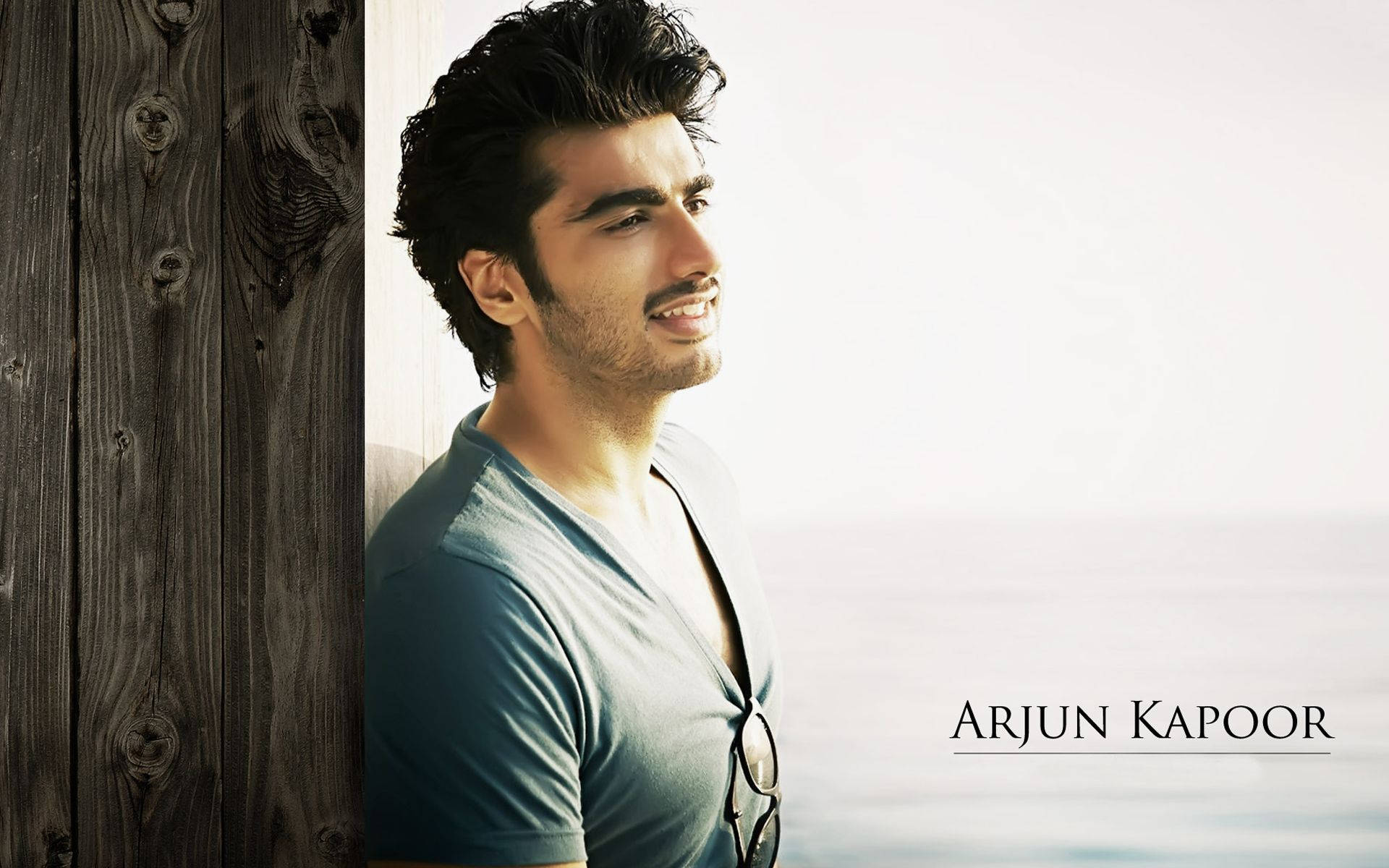 Arjun Kapoor 桌布 壁紙 — 一個深色頭髮、鬍渣的男人靠著明亮的窗戶旁邊的風化木牆，溫柔地微笑。