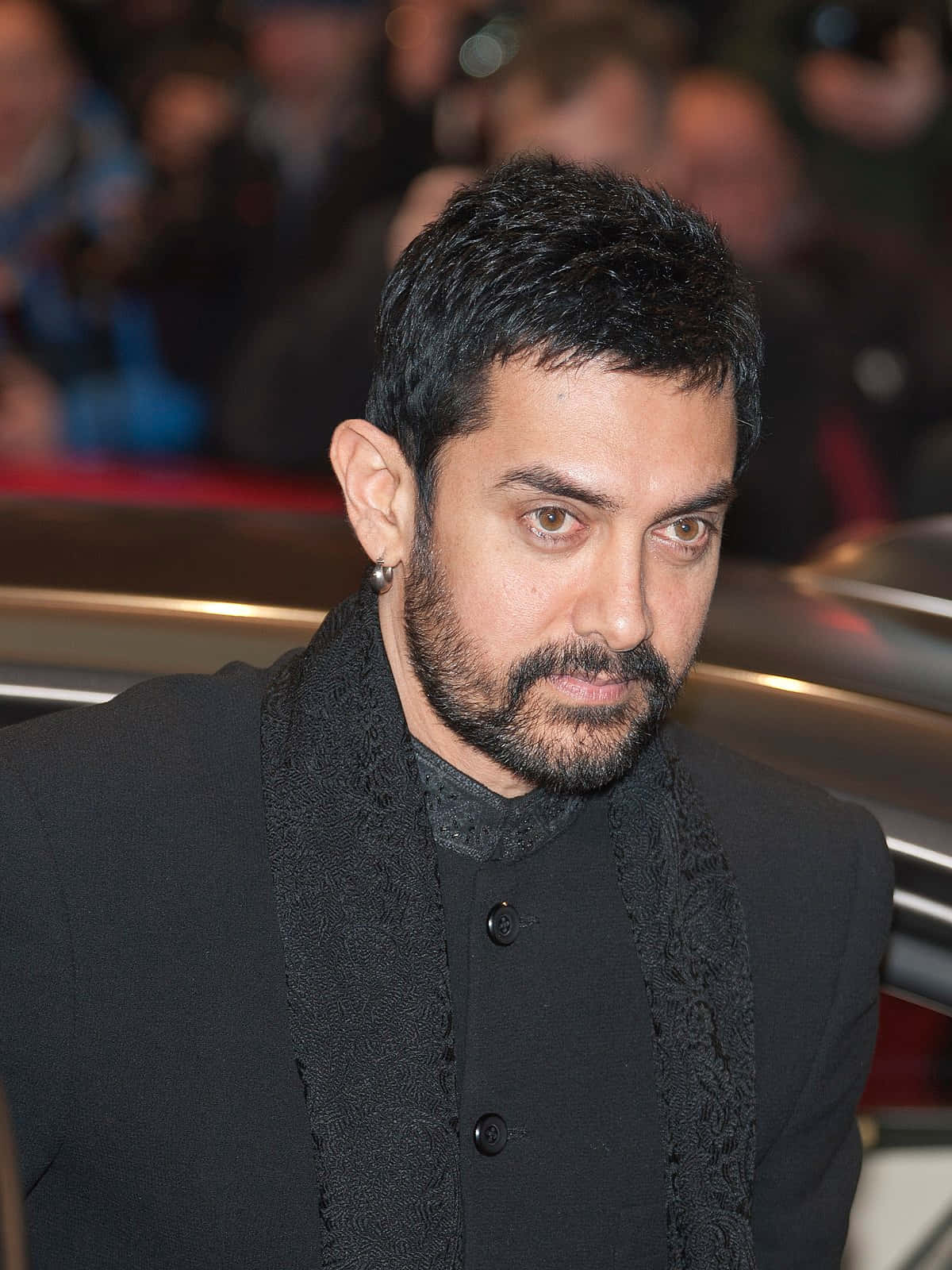 Aamir Khan 桌布 壁紙 — 一個深色頭髮和鬍鬚的男人穿著黑色服裝，在模糊的人群背景下專注地看著相機。