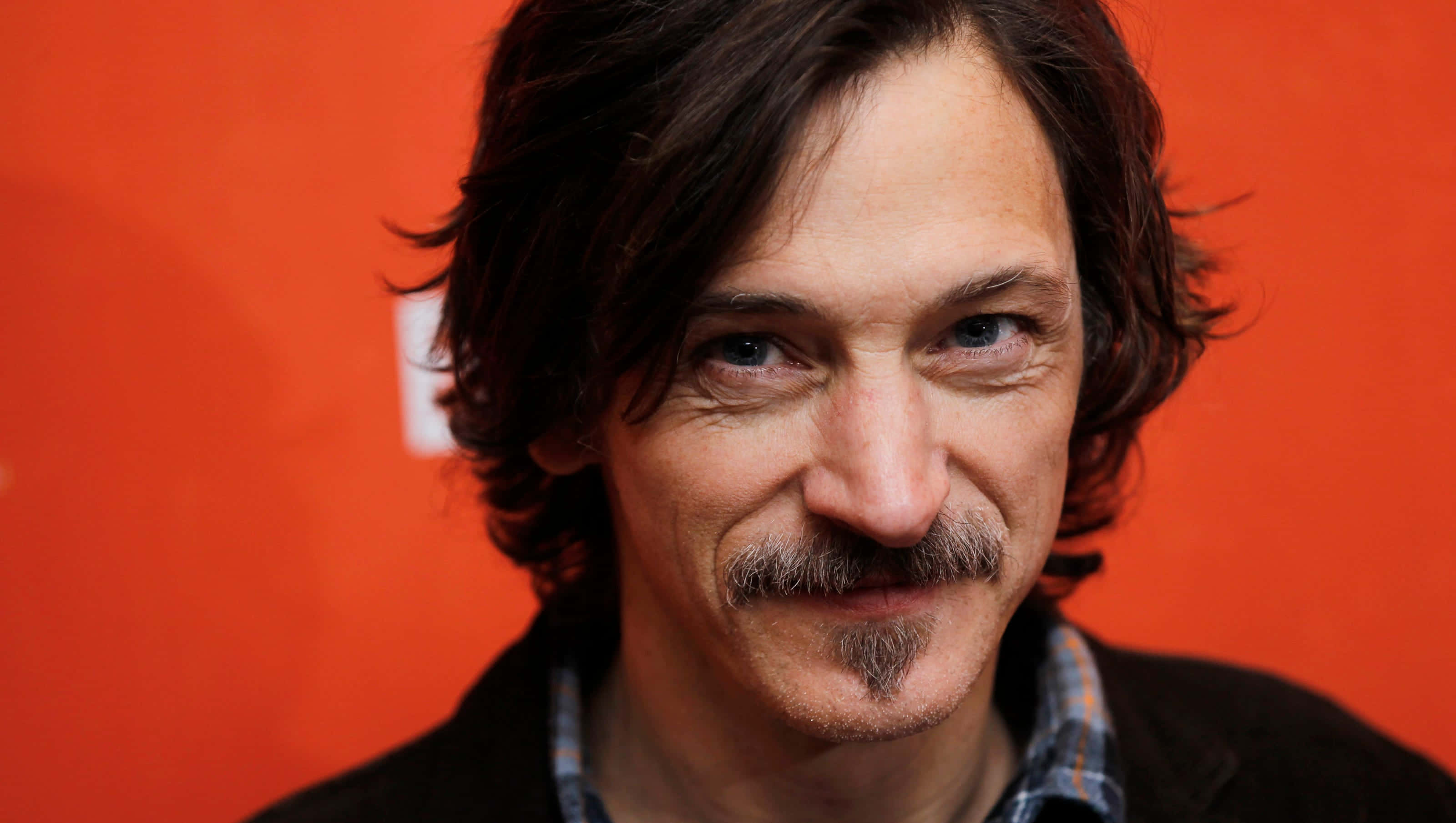 John Hawkes 桌布 壁紙 — 一個深色頭髮和鬍鬚的男子在鮮豔的橙色背景前微笑的特寫肖像。