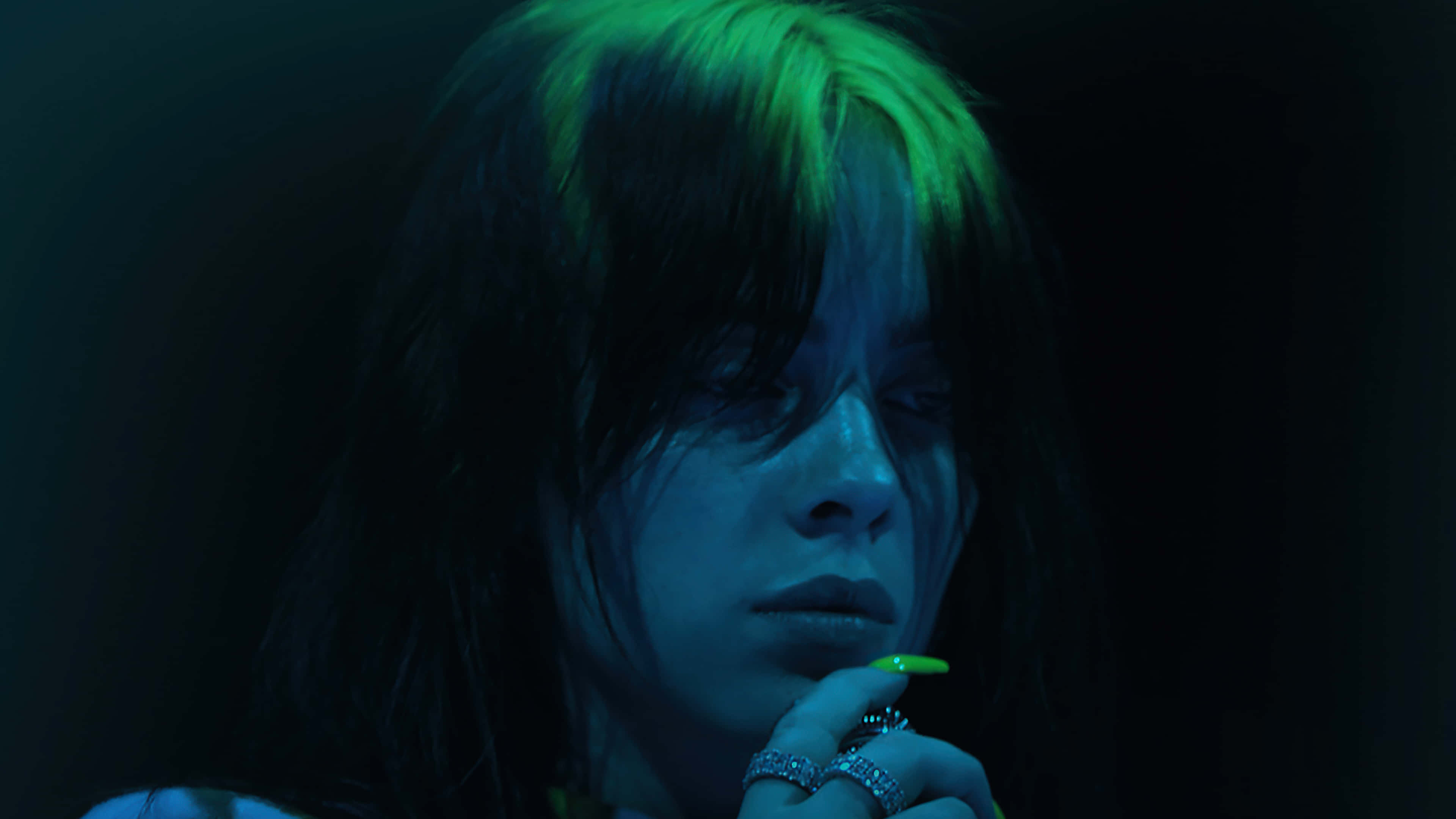 Billie Eilish Laptop 桌布 壁紙 — 一個深色頭髮帶有螢光綠色挑染的人，在憂鬱的藍色燈光下，手靠近臉部，專注地凝視。