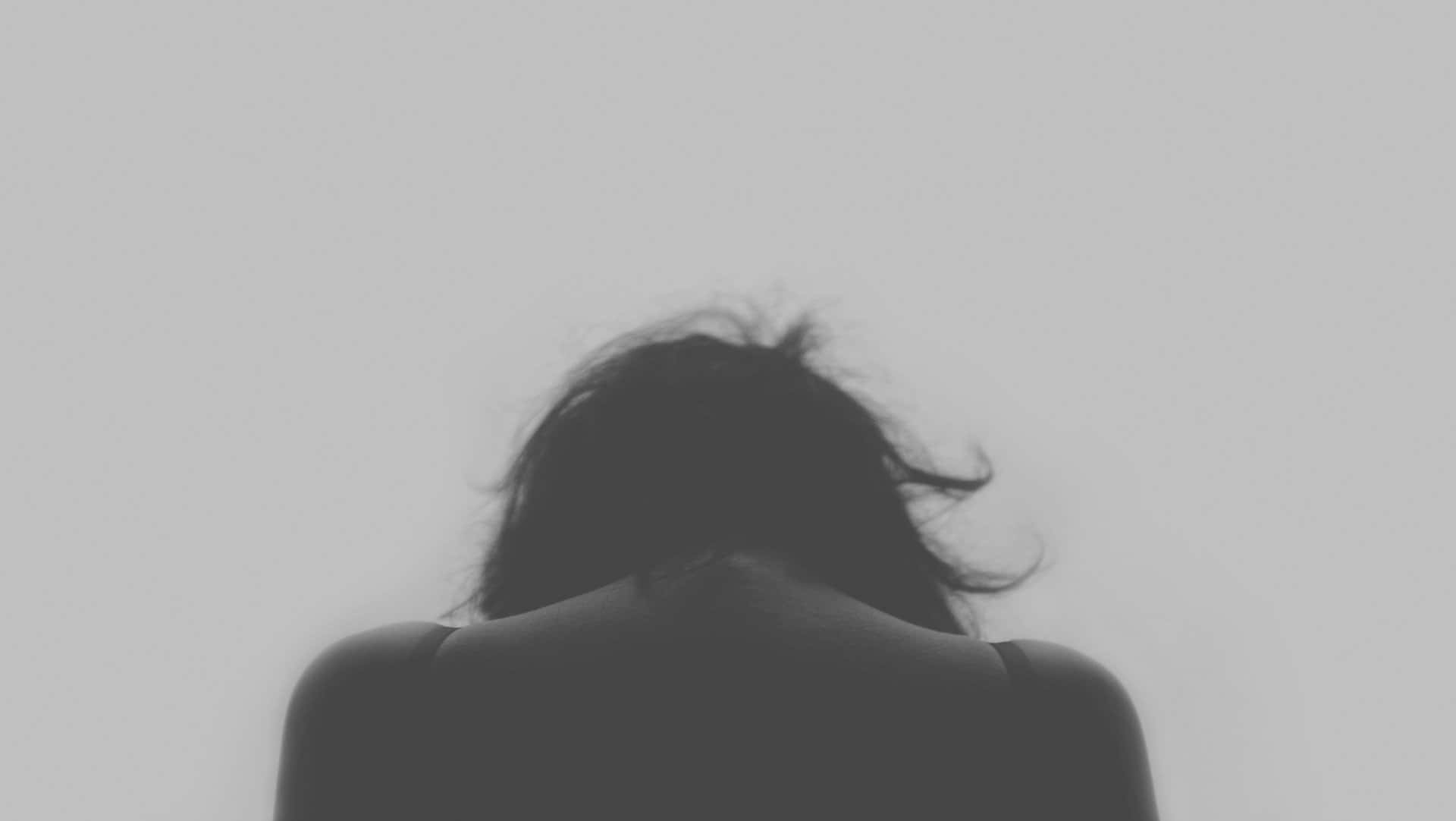 Depression Girl 桌布 壁紙 — 一個深色頭髮的人在單色照片中，將頭低下朝向純淺灰色背景。