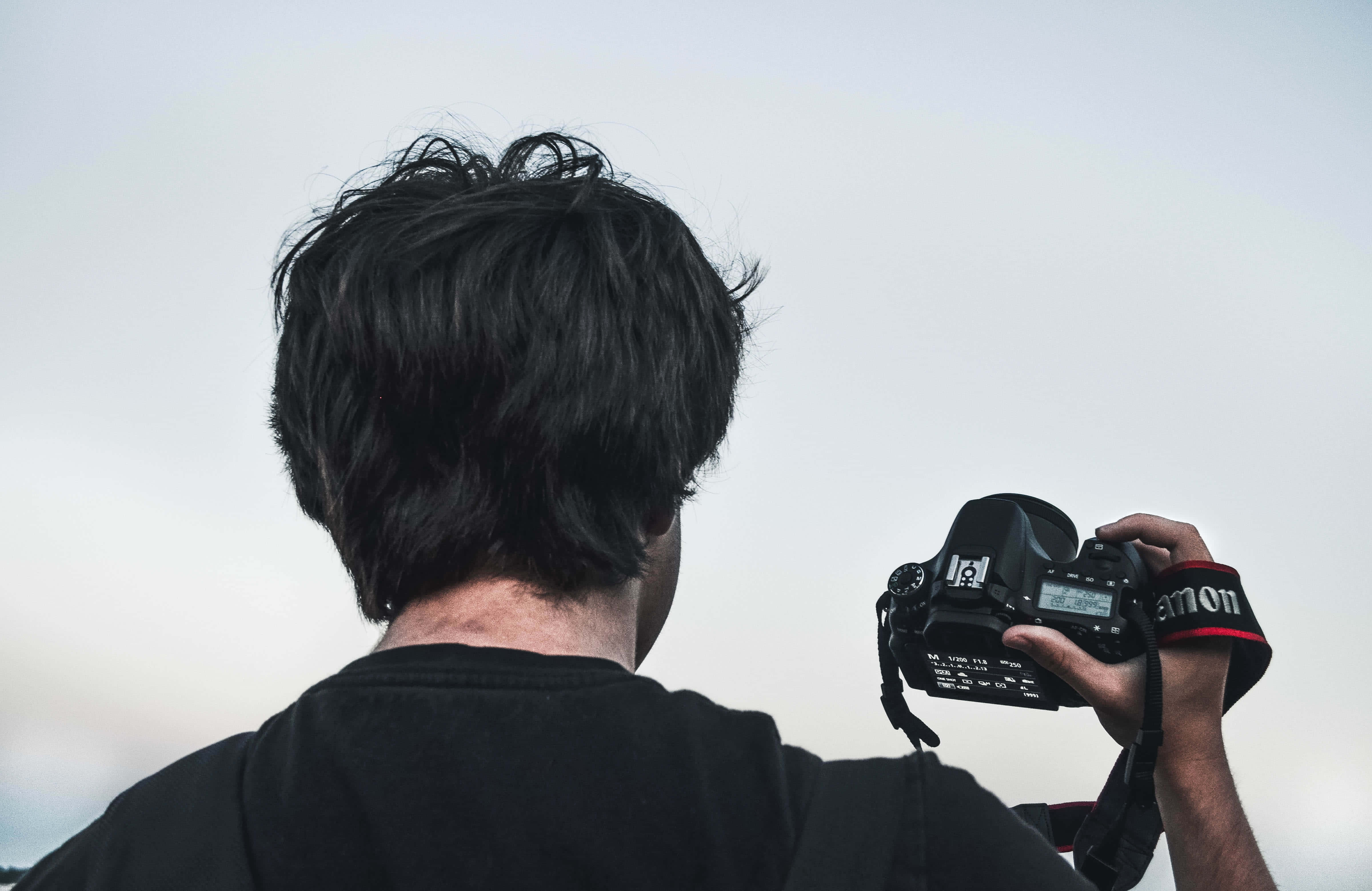 Dslr 桌布 壁紙 — 一個深色頭髮的人舉起黑色Canon DSLR相機，對著蒼白的天空捕捉場景。