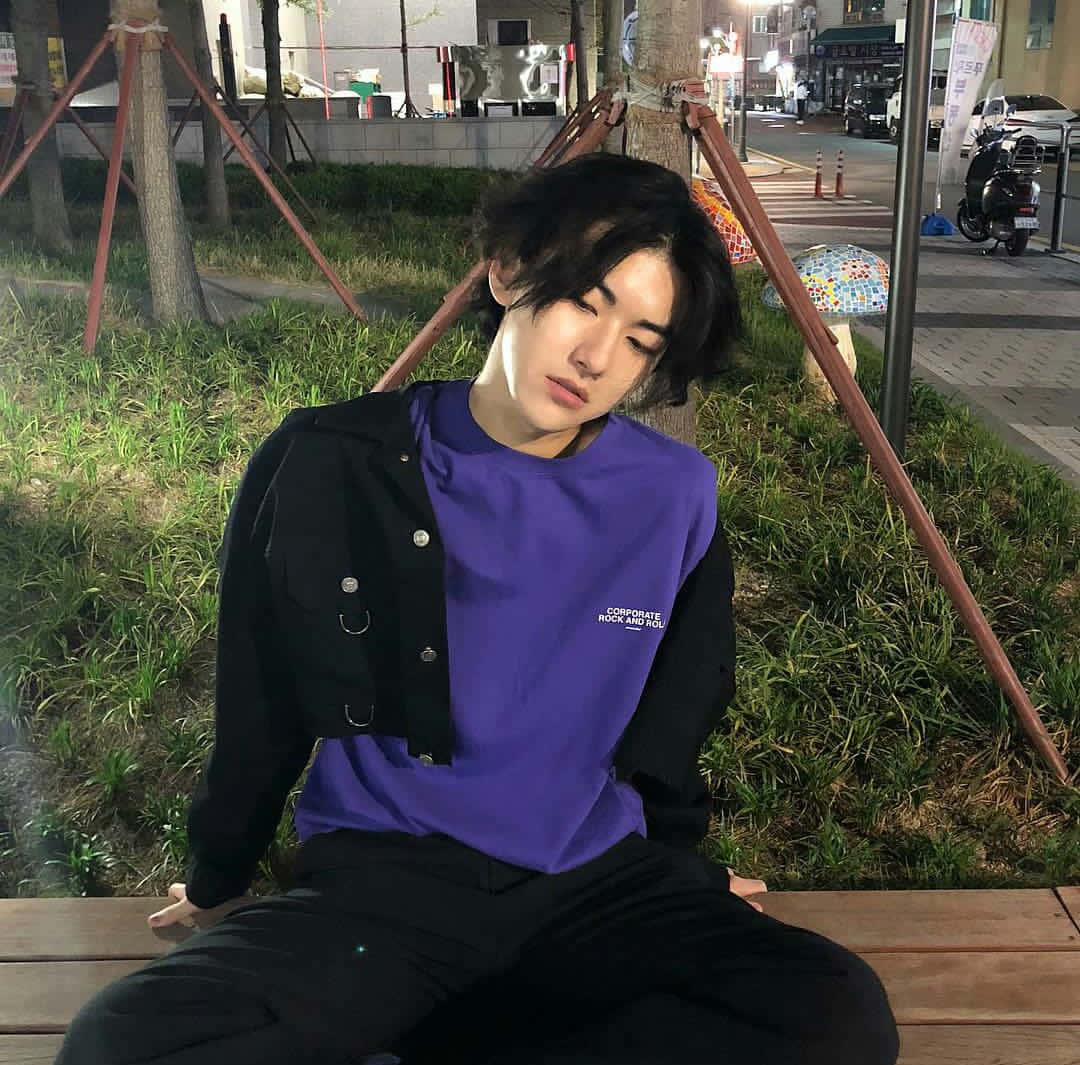 美少年動態圖 桌布 壁紙 — 一個深色頭髮的年輕男子晚上坐在木長椅上，穿著紫色襯衫和黑色外套，被草地和城市街燈包圍。