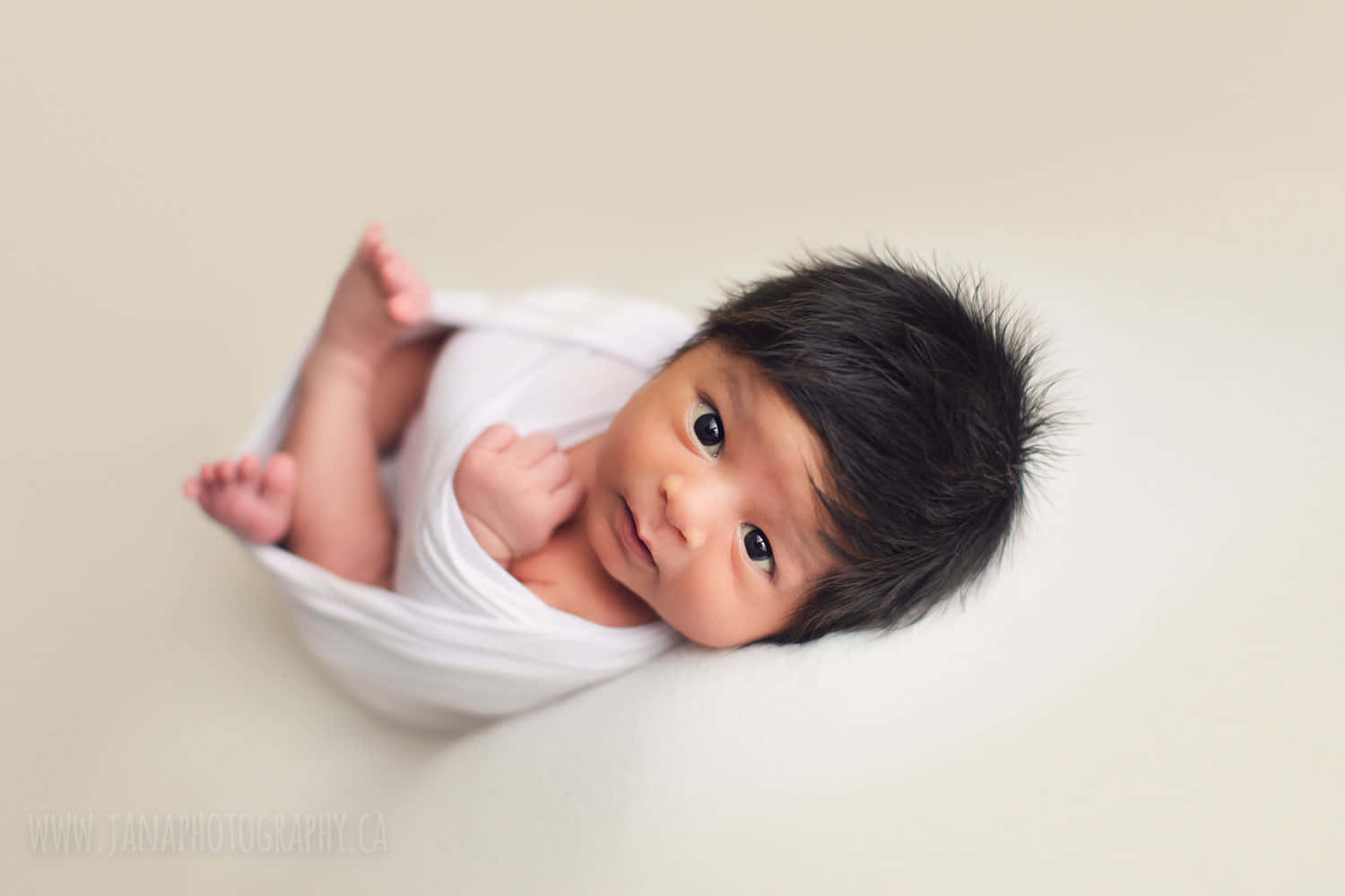 Newborn Baby 桌布 壁紙 — 一個深色頭髮的新生嬰兒用白布包裹著，躺在柔軟的米色表面上，睜著大大的好奇眼睛向上凝視。