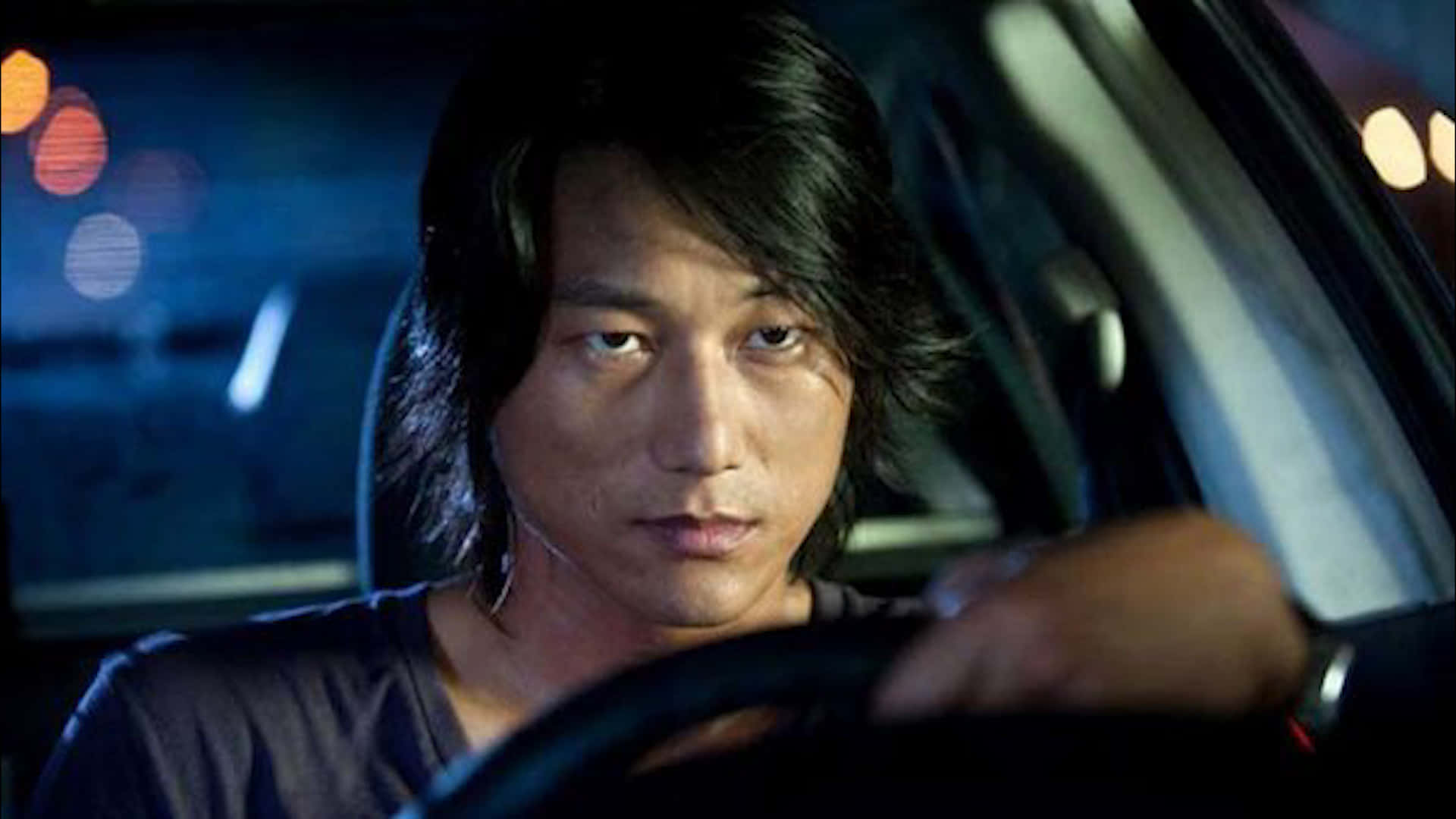 Fast And Furious 桌布 壁紙 — 一個深色頭髮的男人在夜間車內握著方向盤，背後有模糊的城市燈光閃爍。