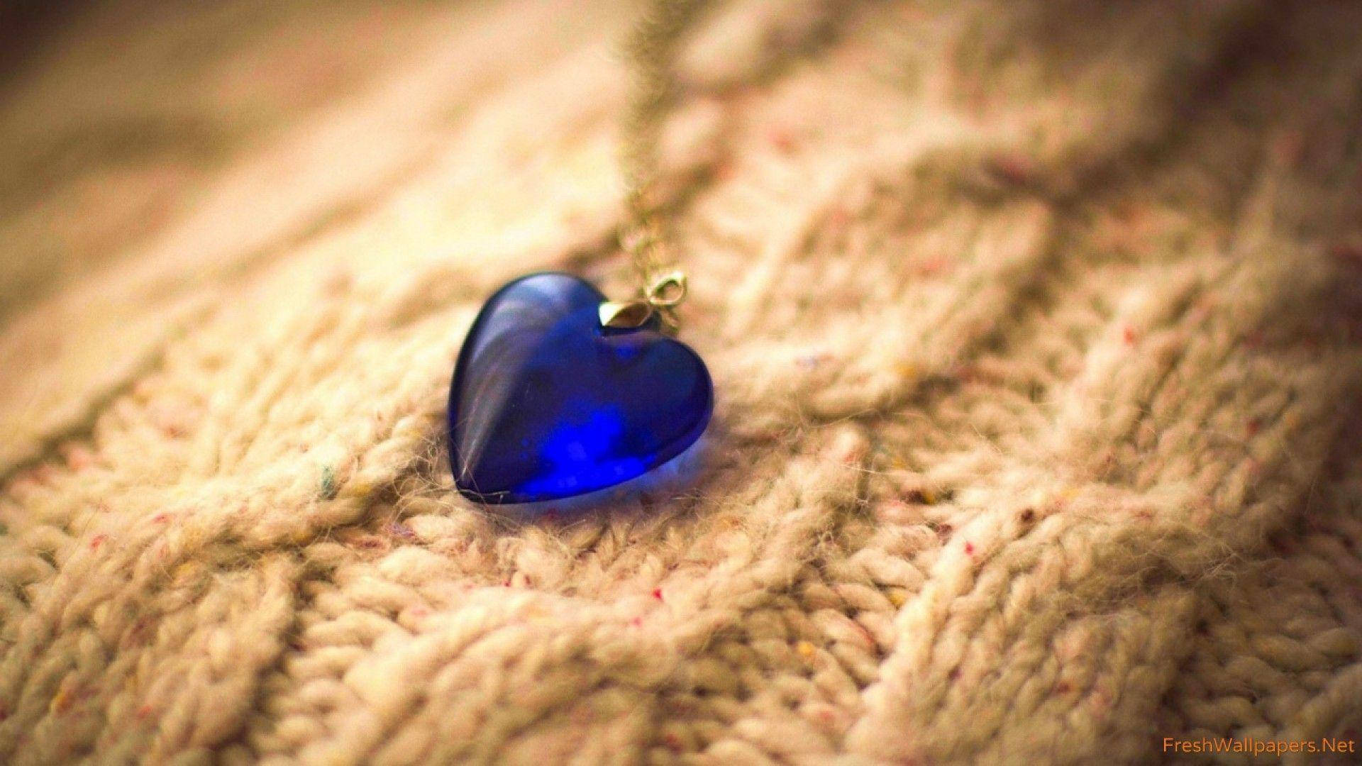Blue Heart 桌布 壁紙 — 一個深藍色心形吊墜放在有紋理的米色針織布料上，帶有柔和溫暖的燈光。