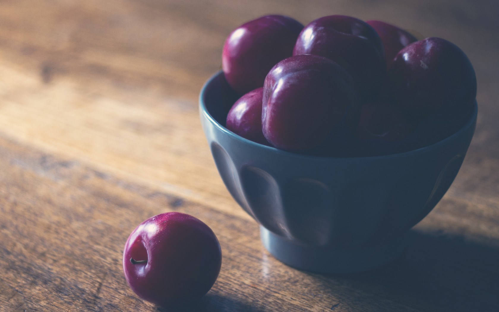 Fruits 桌布 壁紙 — 一個深藍色的碗裝滿了光澤的紫色李子，放在溫暖的木桌上，旁邊還有一個李子。