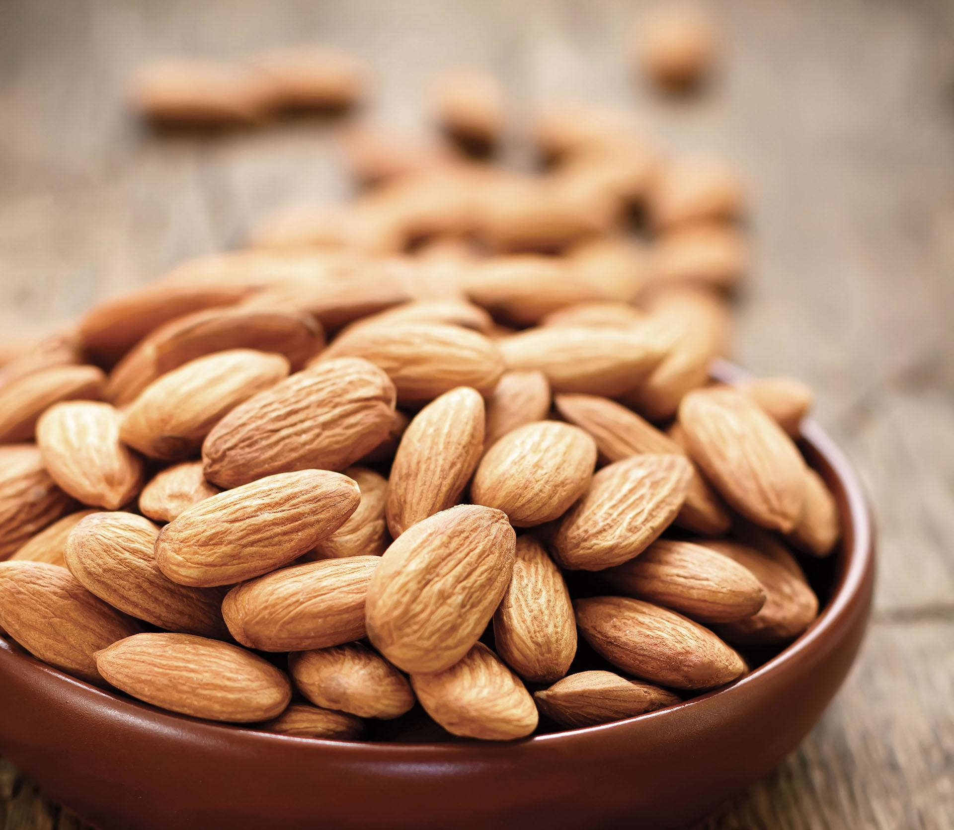 Almond 桌布 壁紙 — 一個淺棕色碗裝滿了有紋理的杏仁，放在質樸的木質表面上，後面散落著堅果。