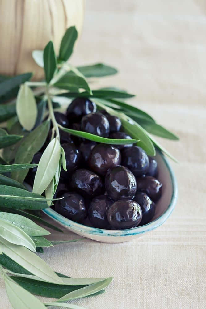 Black Olives 桌布 壁紙 — 一個淺碗盛裝著光澤的黑橄欖，放在有紋理的米色布上，旁邊有新鮮的綠色橄欖葉。