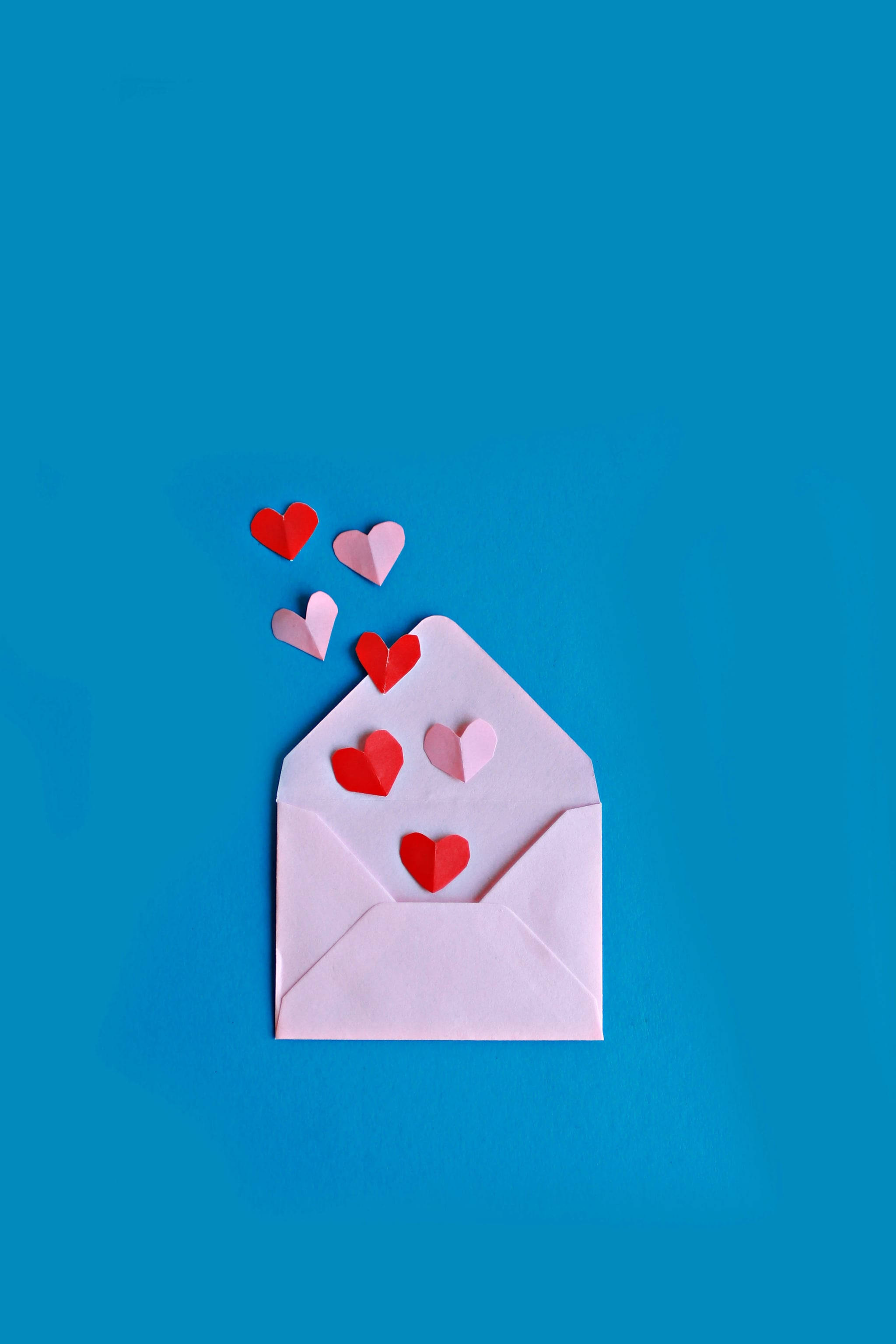 cute iPhone lock screen 桌布 壁紙 — 一個淺粉紅色信封放在鮮豔的藍色背景上，紅色和粉紅色的紙愛心從信封中飄出。