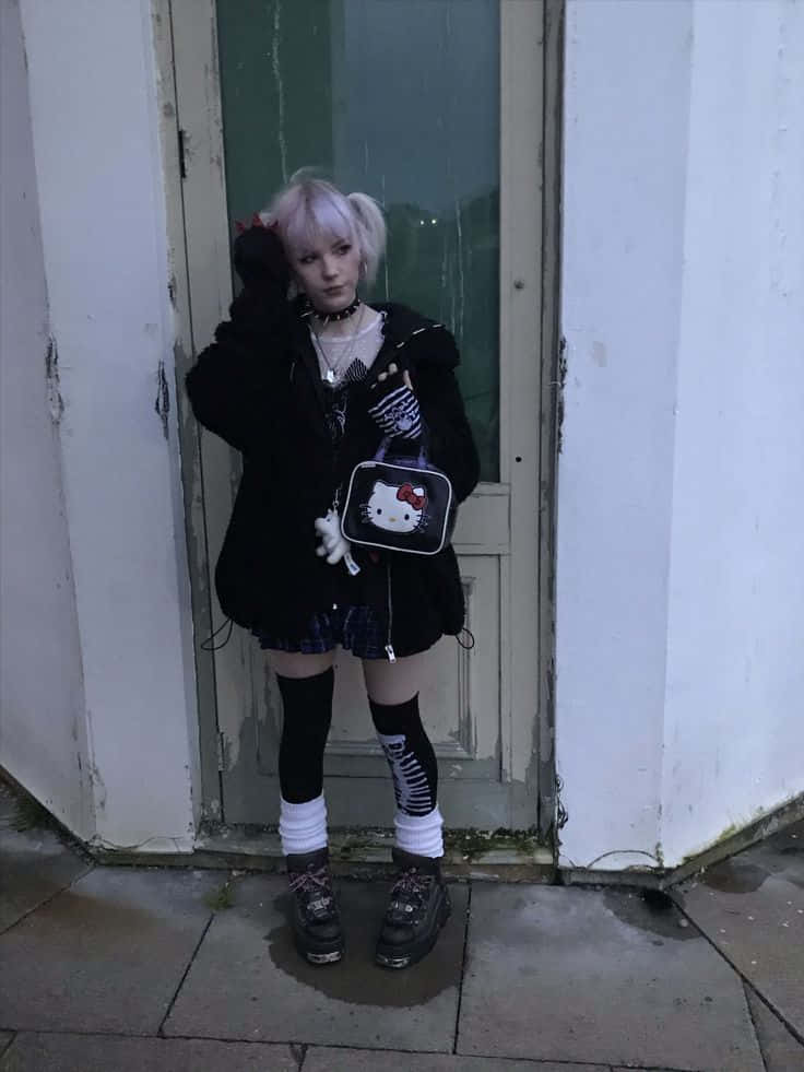 emo 美學 桌布 壁紙 — 一個淺色頭髮的人穿著哥德蘿莉塔服裝，站在一扇風化的門旁，拿著一個Hello Kitty手提包。