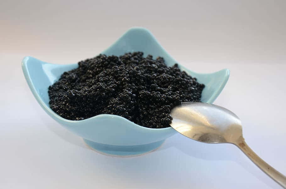 Utensils 桌布 壁紙 — 一個淺藍色扇貝形碗盛載著閃閃發光的黑魚子醬，旁邊有一把銀湯匙放在白色表面上。