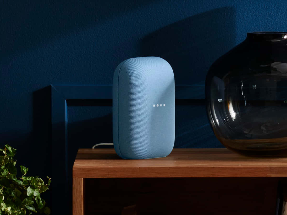google 桌布 壁紙 — 一個淺藍色智慧音箱帶有發光點，放在木架上，旁邊有一個深色玻璃花瓶和綠色植物，背景是深藍色牆壁。
