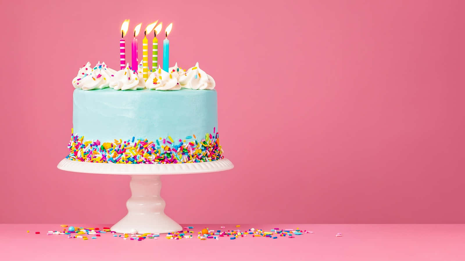 Birthday Cake 桌布 壁紙 — 一個淺藍色生日蛋糕，上面有彩色糖粒和五根點燃的蠟燭，放在白色支架上，背景是純粉紅色。