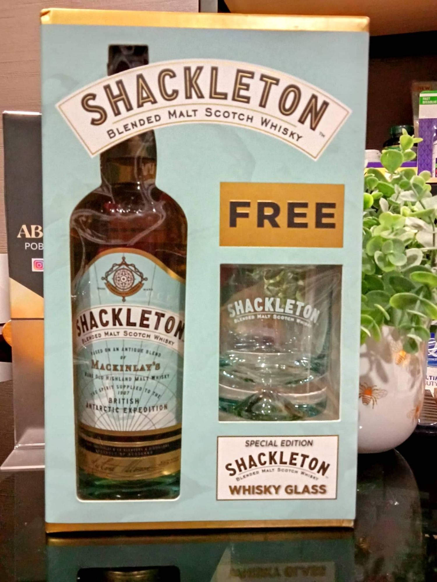 Drink 桌布 壁紙 — 一個淺藍色盒子展示一瓶Shackleton威士忌和一個免費威士忌杯，旁邊有一棵綠色植物。