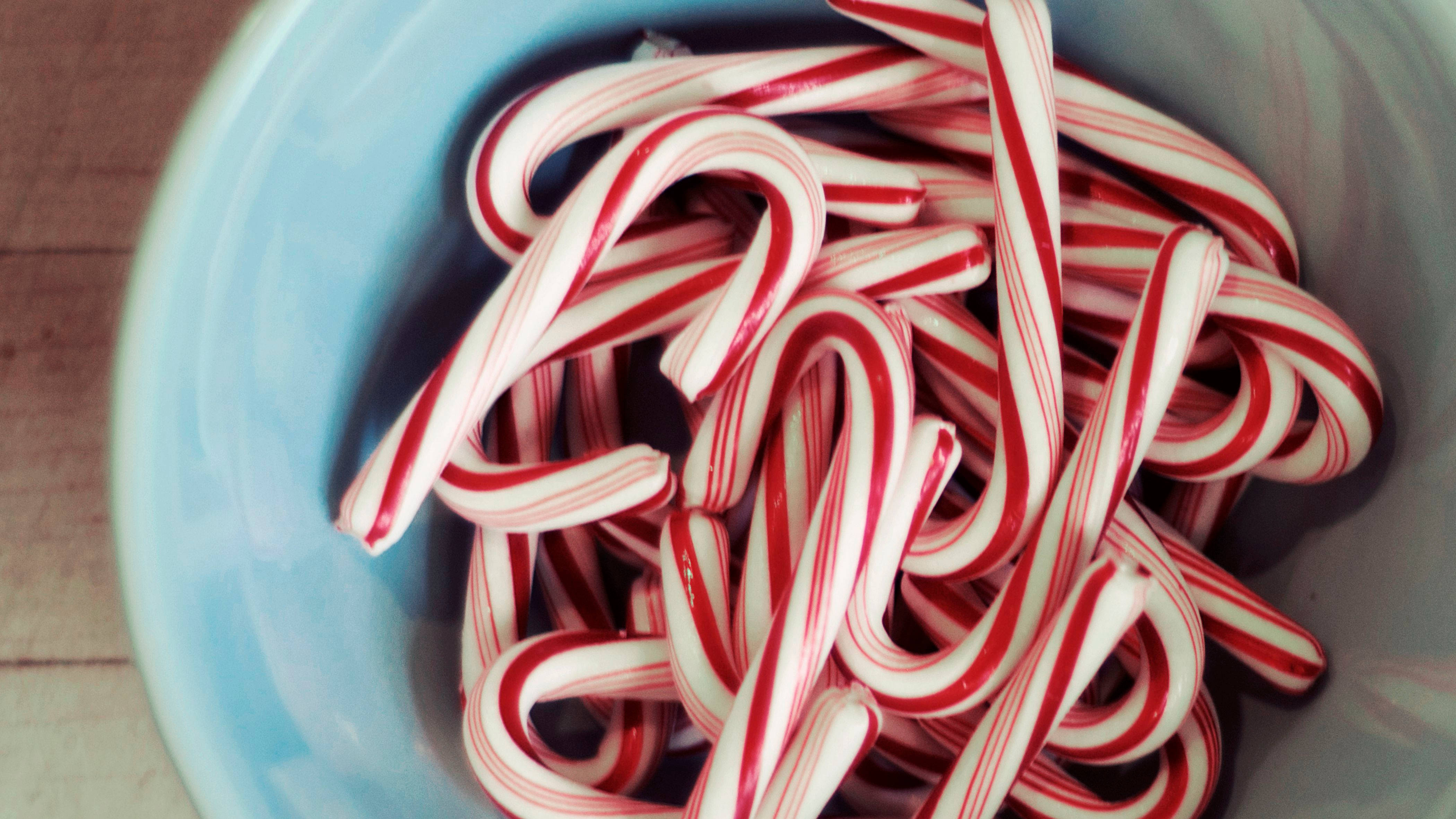 Candy Cane 桌布 壁紙 — 一個淺藍色碗，裝滿經典紅白條紋拐杖糖，放在木質表面上。