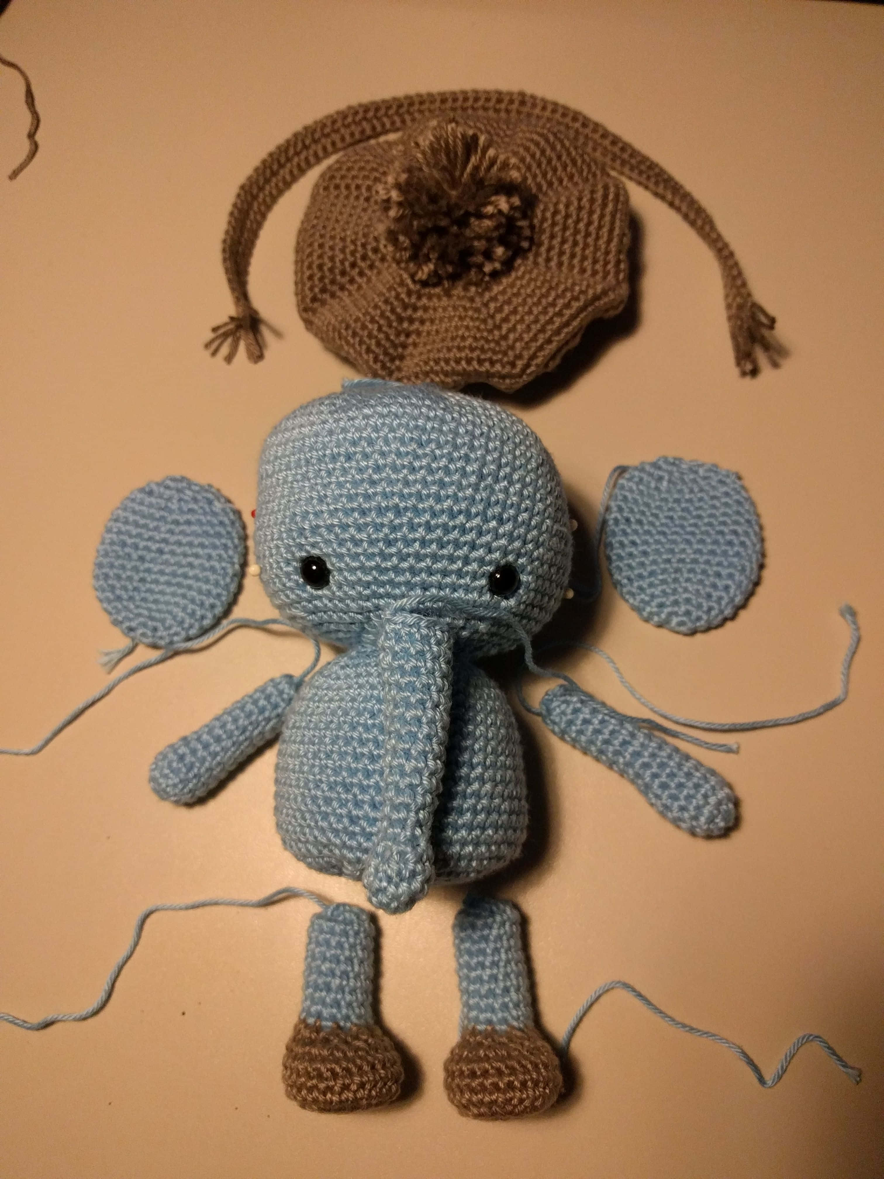 Emily Elephant 桌布 壁紙 — 一個淺藍色鉤針大象玩具，配有棕色腳和相配的帽子，被拆解後放在米色表面上。