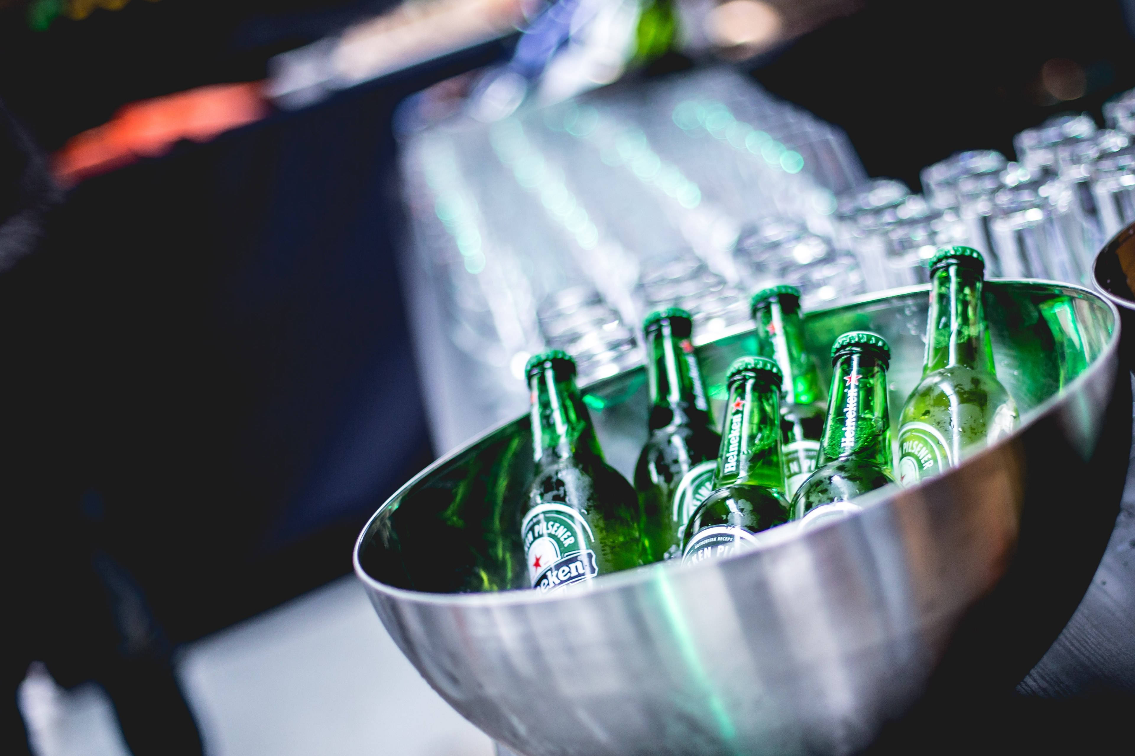 alcohol 桌布 壁紙 — 一個淺金屬碗裝滿冰涼的綠色海尼根啤酒瓶，旁邊放著疊起的透明玻璃杯，在燈光昏暗的酒吧環境中。