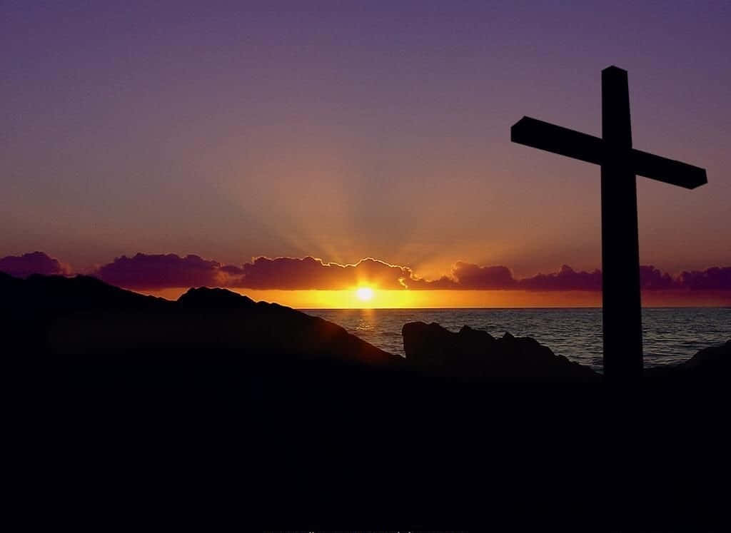 Holy 桌布 壁紙 — 一個清晰的十字架剪影矗立在岩石海岸上，夕陽在海面上落下，穿過紫色雲層投射出溫暖的金色光芒。