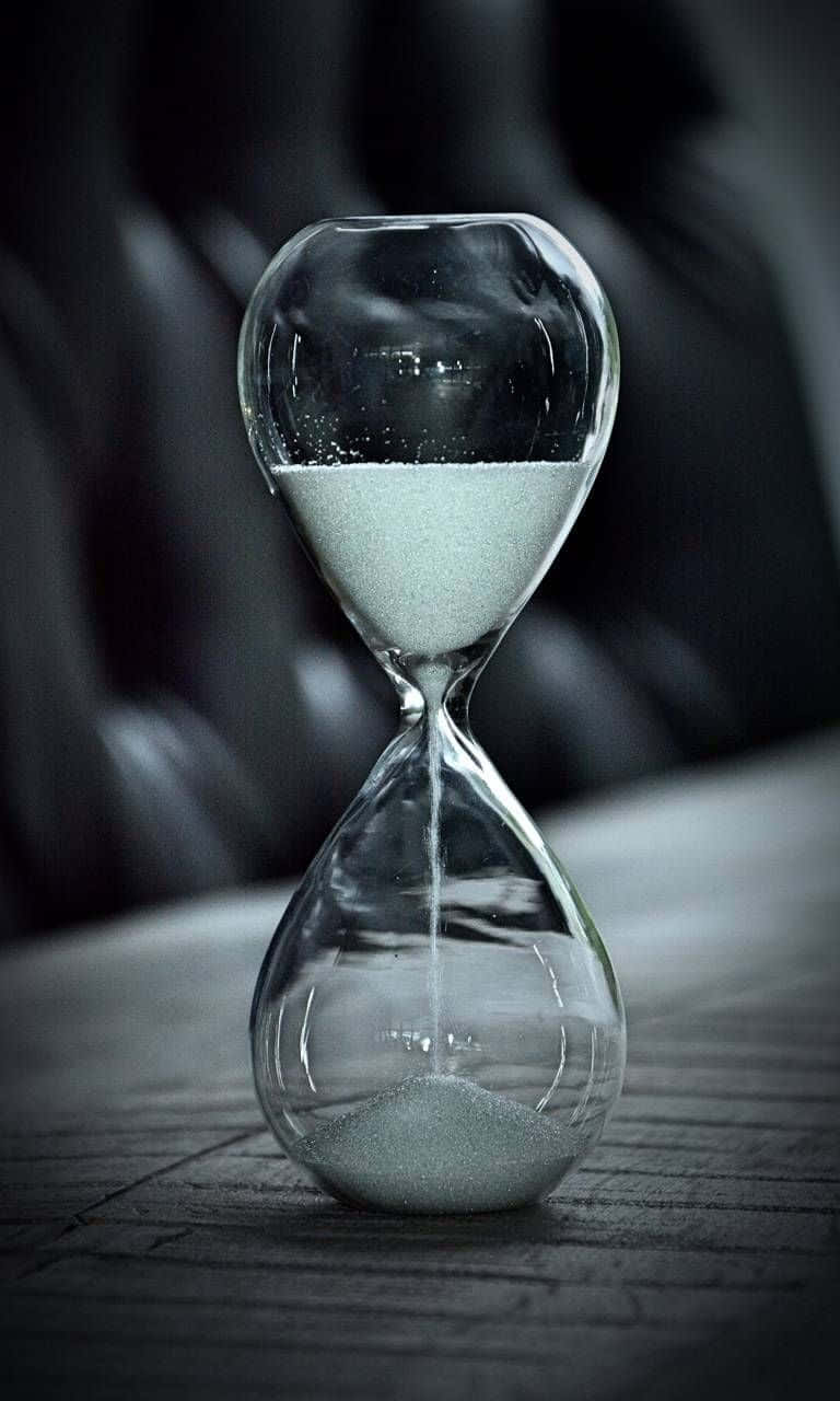 Time 桌布 壁紙 — 一個清晰的沙漏，白色沙子穩定流動，放置在木質表面上，背景是深色皮革。