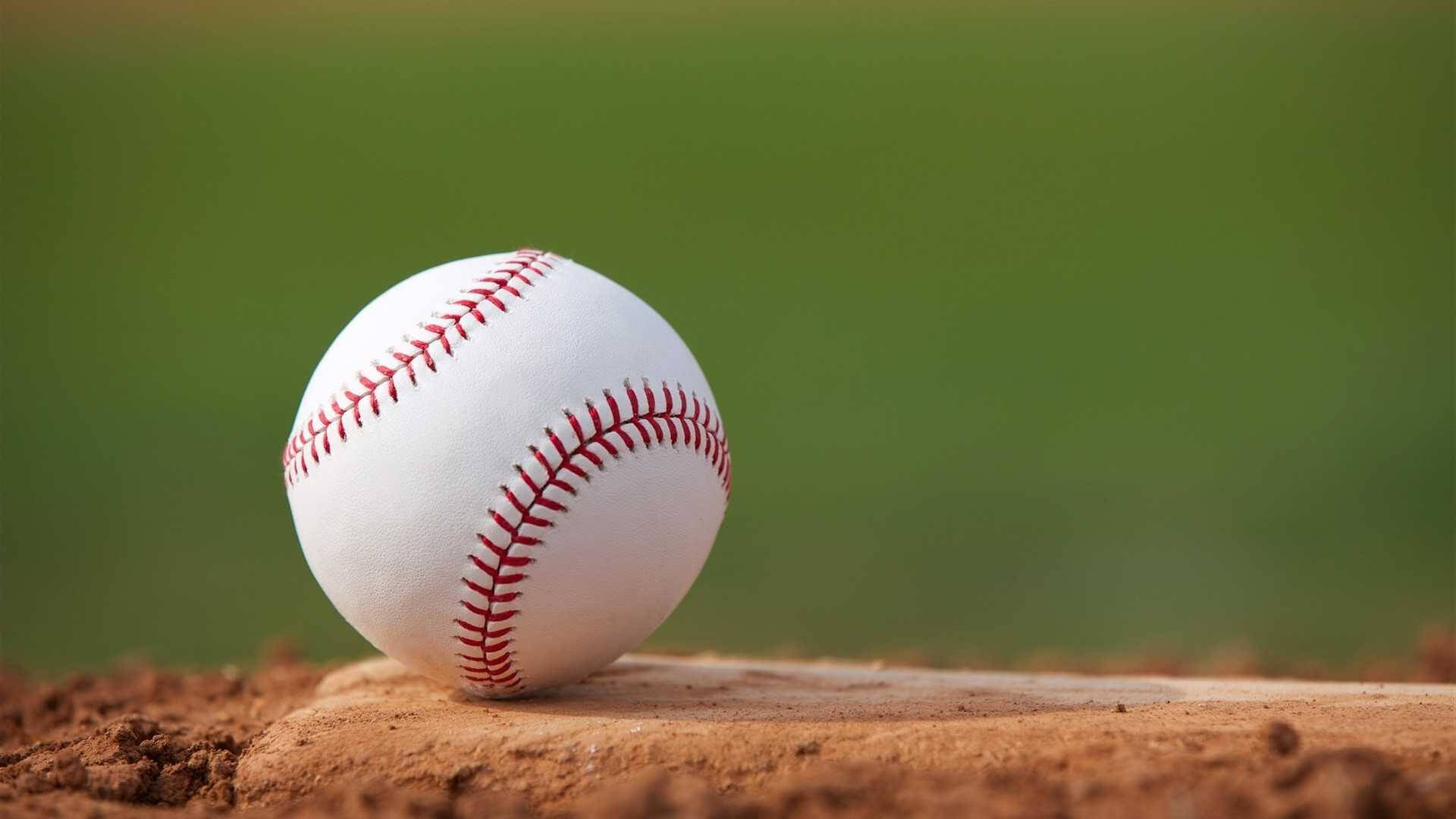 Cute Softball 桌布 壁紙 — 一個清晰的白色棒球，帶有紅色縫線，放置在磨損的木製底座上，周圍是棕色泥土，背景是模糊的綠色球場。