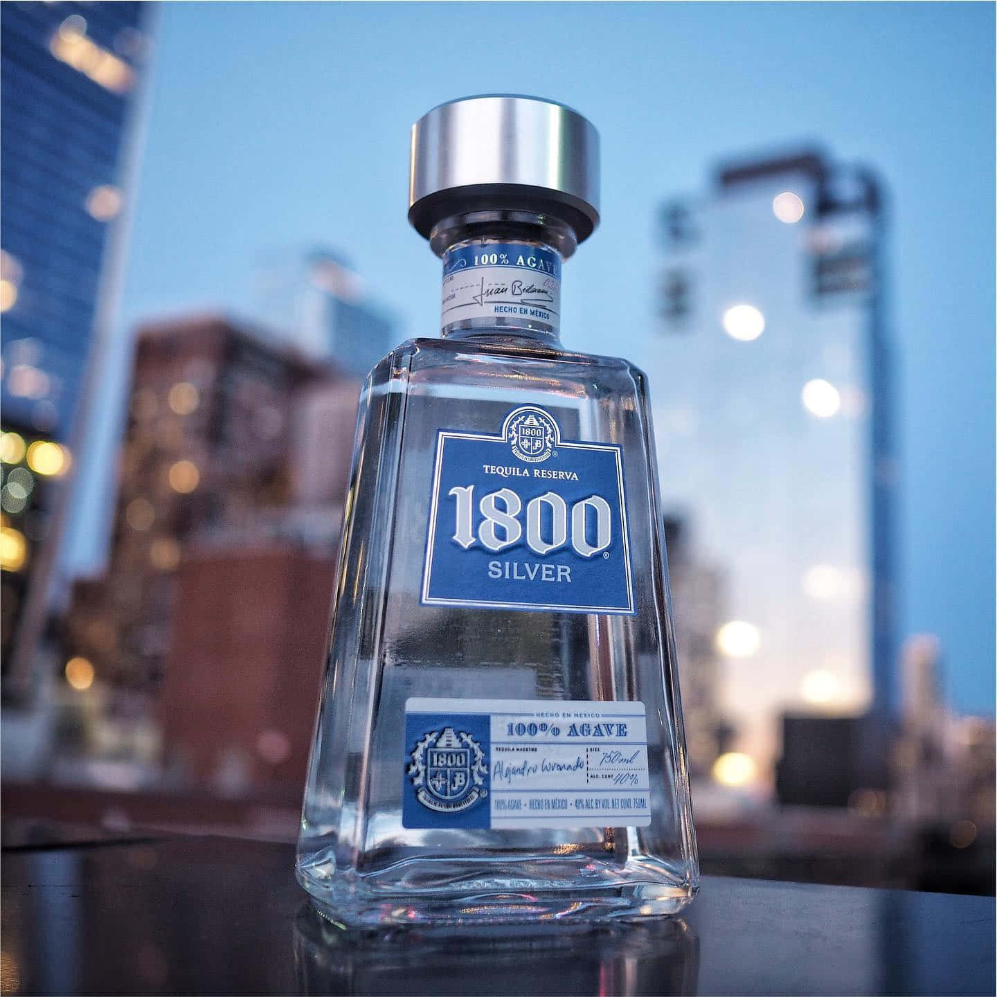 alcohol 桌布 壁紙 — 一個清澈的1800 Silver龍舌蘭酒瓶在屋頂邊緣閃耀，背景是暮色中柔和模糊的城市天際線。