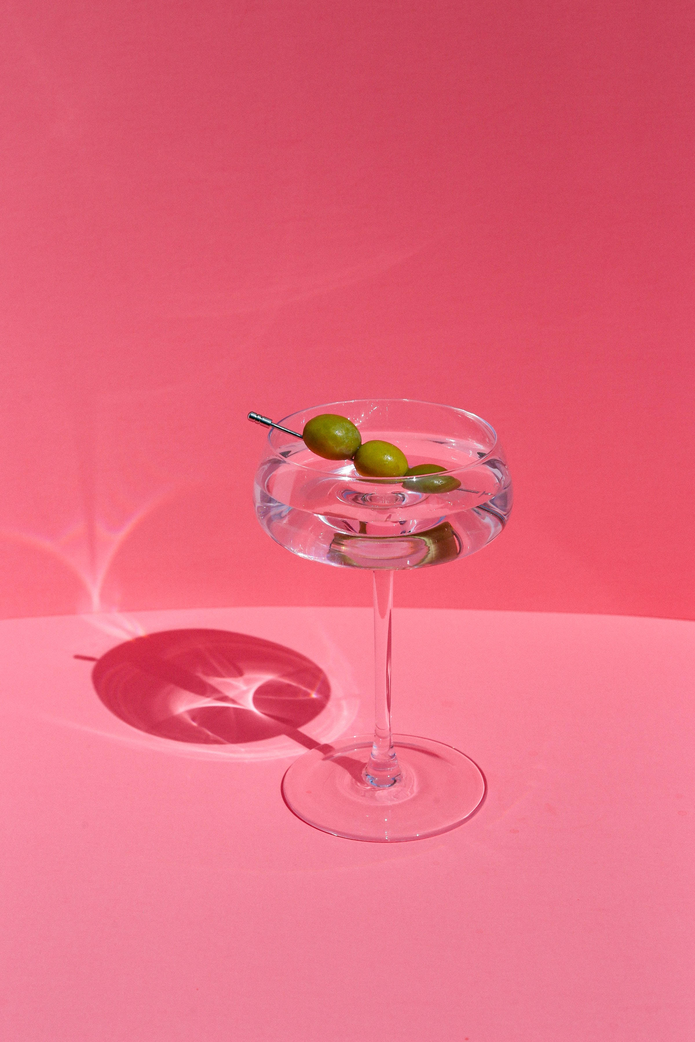 drinks 桌布 壁紙 — 一個清澈的馬丁尼酒杯，杯中有三顆青橄欖插在牙籤上，放在充滿活力的粉紅色背景前，投射出清晰的陰影。