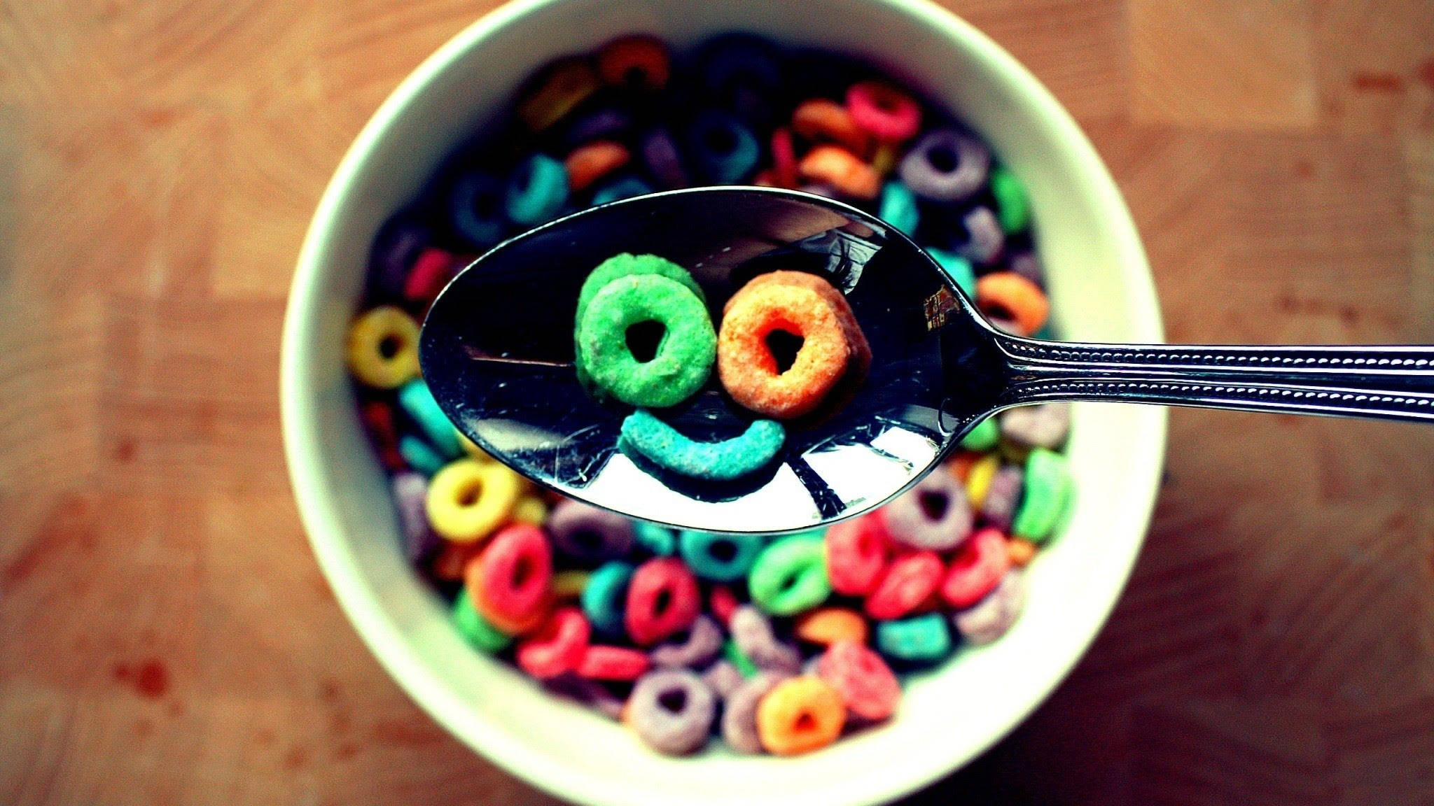 Happy Face 桌布 壁紙 — 一個湯匙上放著兩個穀物圈，形成一個笑臉，放在木桌上一個色彩繽紛的穀物碗上方。