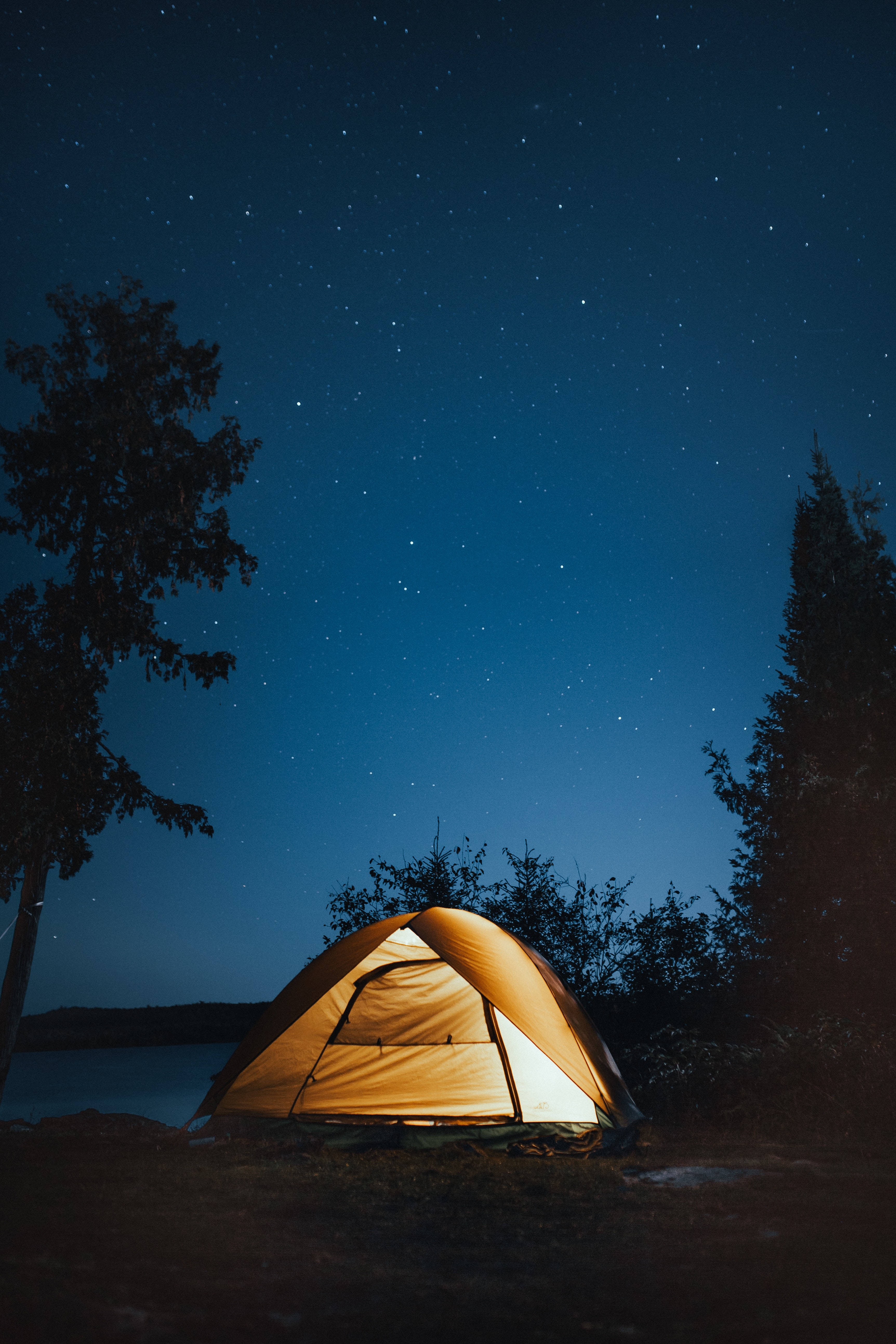 夜空 桌布 壁紙 — 一個溫暖照亮的橙色帳篷在廣闊的星空下，在平靜的湖邊，被黑暗的樹木環繞著發光。