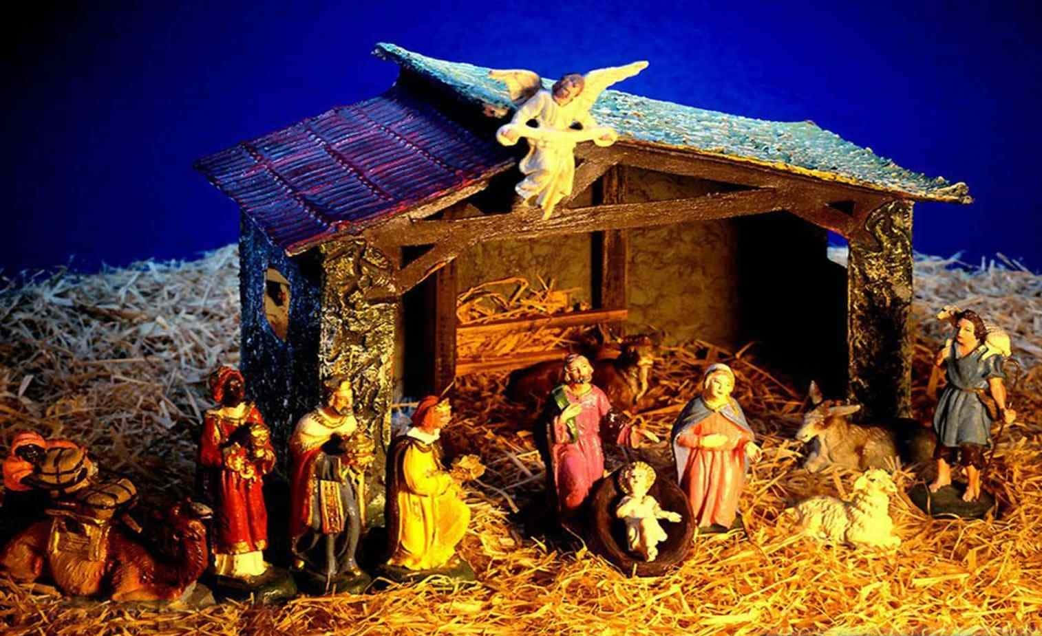 Nativity 桌布 壁紙 — 一個溫暖照亮的耶穌誕生場景，有瑪利亞、約瑟、嬰兒耶穌、牧羊人和動物的雕像，在藍天下有一位天使在上方。