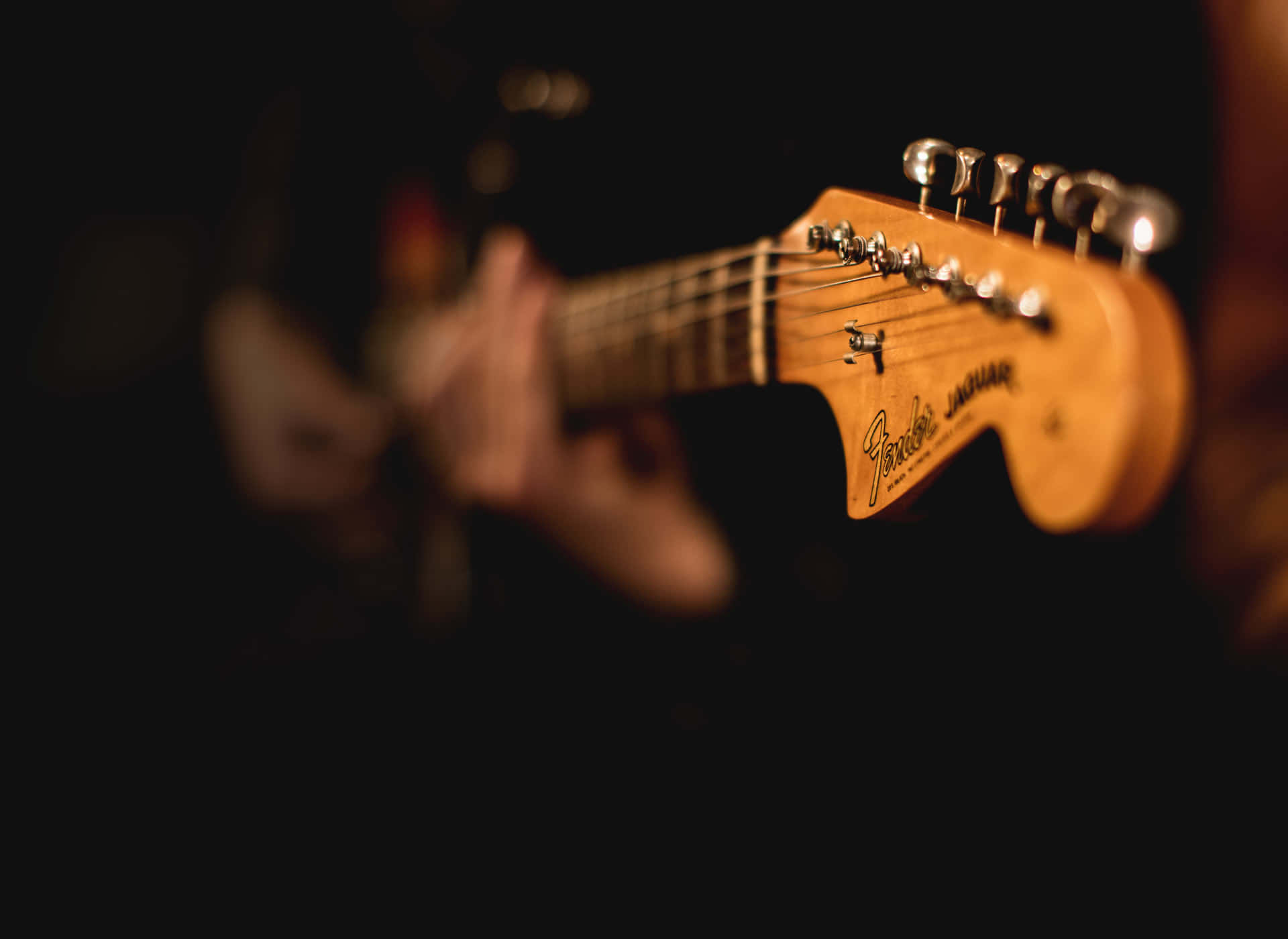 音樂 桌布 壁紙 — 一個溫暖照明的Fender Jaguar吉他琴頭，在黑暗模糊的背景中清晰對焦，有一隻手在彈奏。