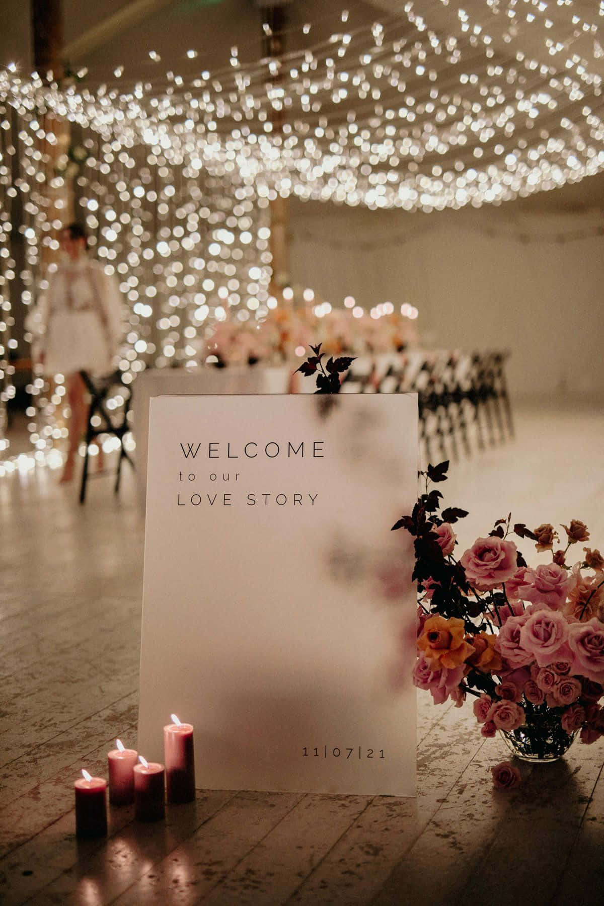Roses Wedding 桌布 壁紙 — 一個溫暖照明的婚禮歡迎牌，周圍環繞著閃爍的仙女燈、蠟燭和粉紅玫瑰，置於木地板上。