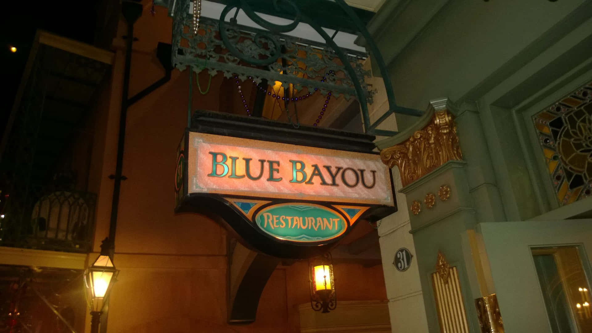 Blue Bayou 桌布 壁紙 — 一個溫暖照明的復古招牌，為Blue Bayou Restaurant在夜間懸掛在帶有華麗金色裝飾和發光燈籠的建築物上。