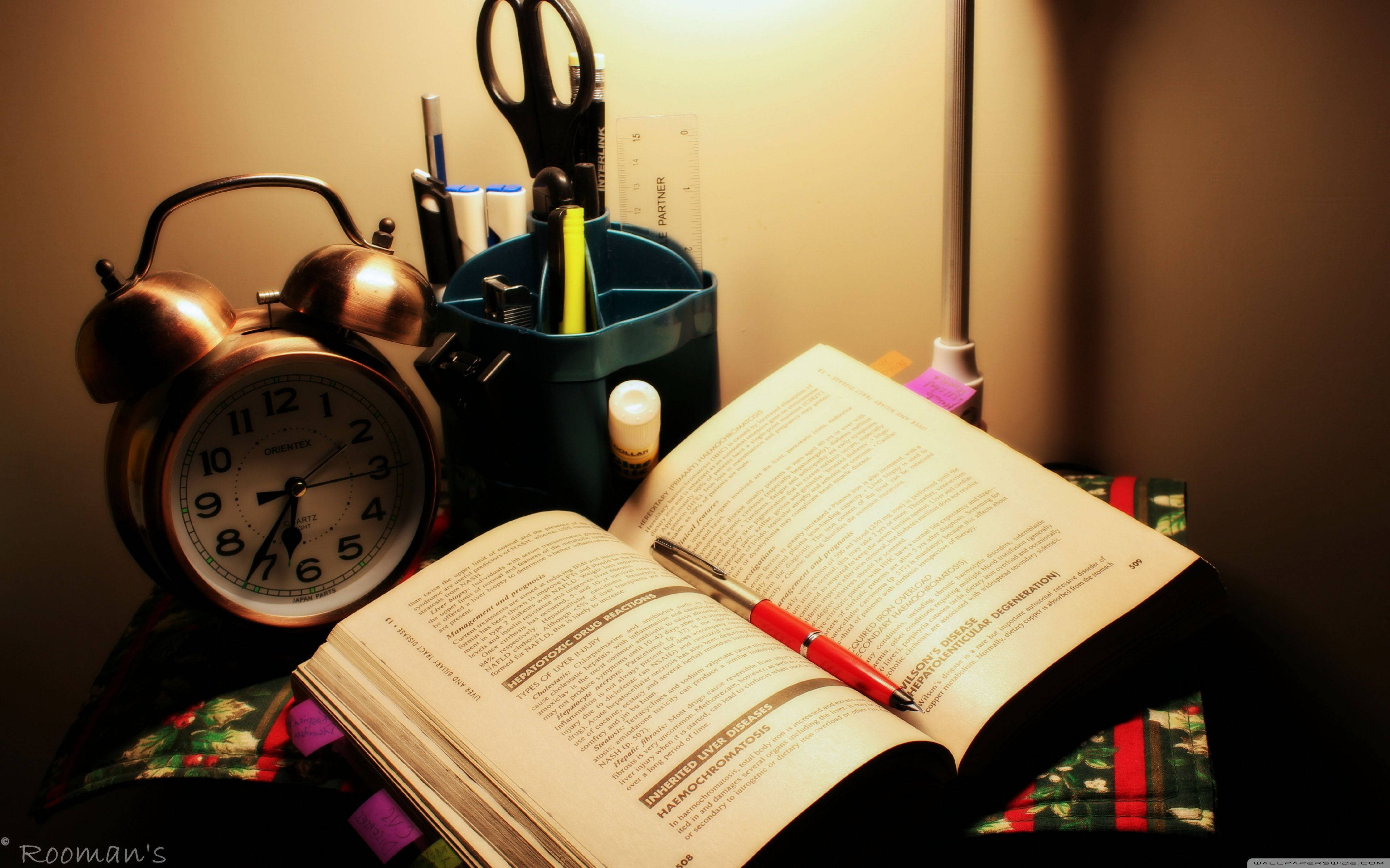 Studying 桌布 壁紙 — 一個溫暖照明的書桌場景，有一本打開的書、紅色筆、鬧鐘和文具在燈光下。