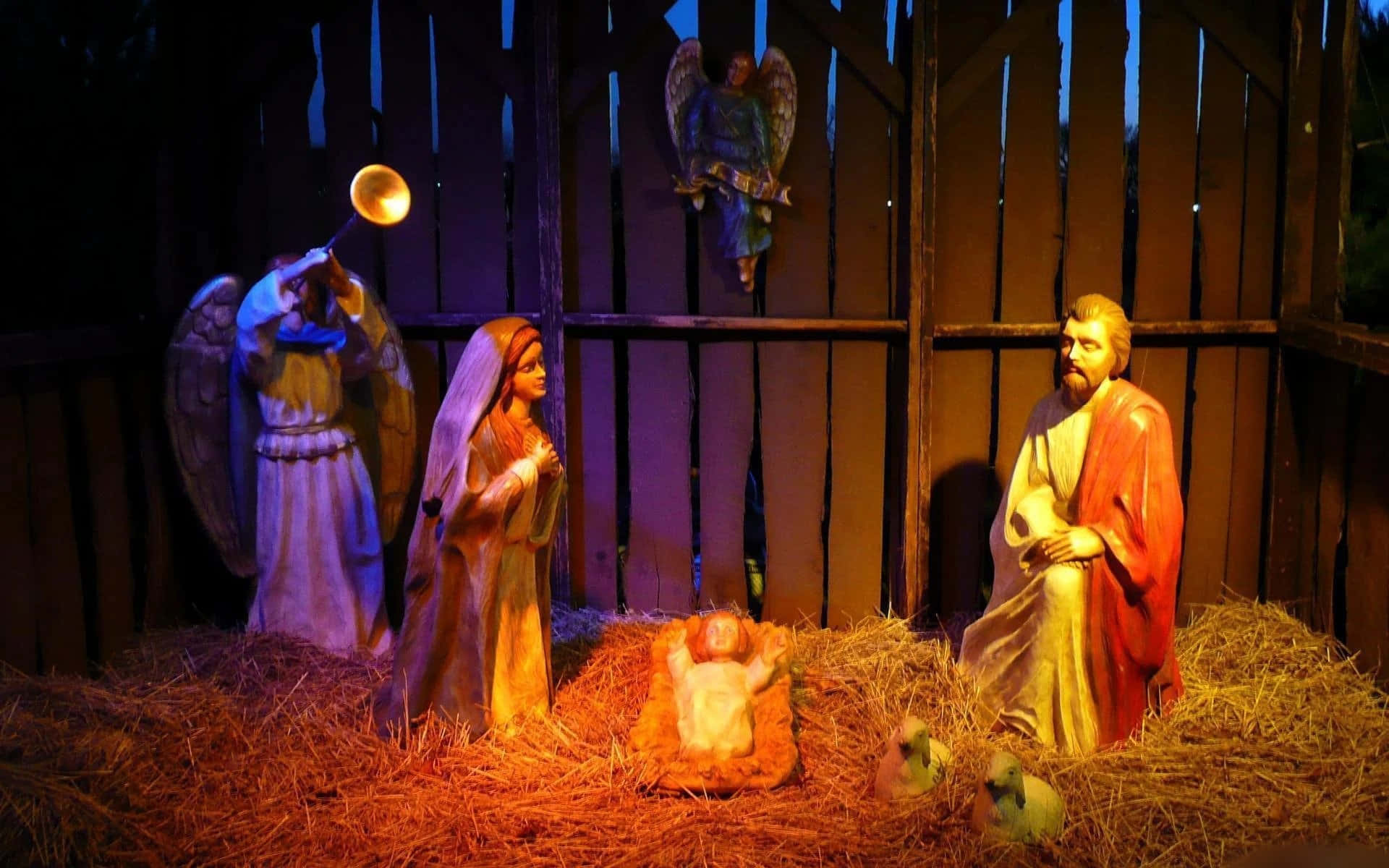 Nativity 桌布 壁紙 — 一個溫暖照明的耶穌誕生場景，有瑪利亞、約瑟、嬰兒耶穌和天使的雕像，位於一個有乾草的木製馬廄中。