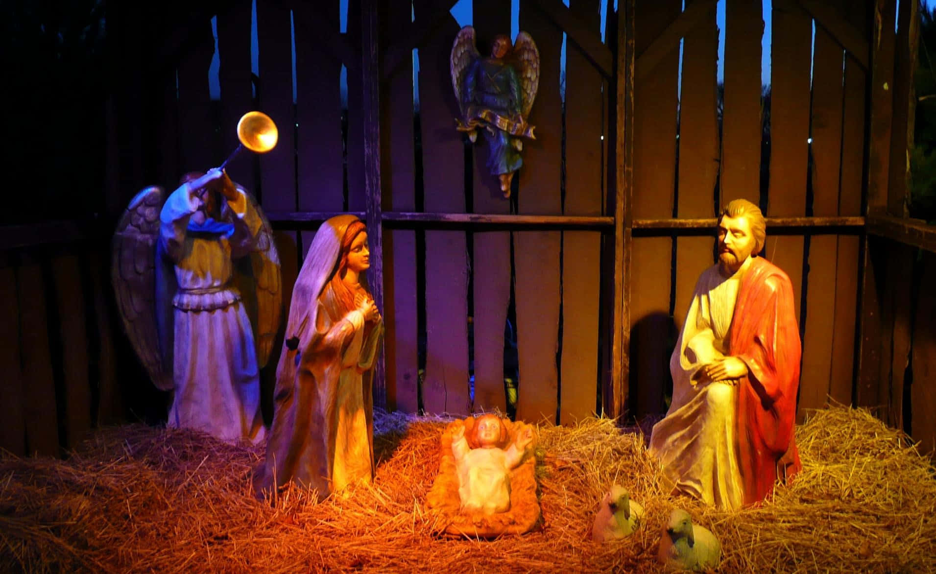 Nativity Scene 桌布 壁紙 — 一個溫暖照明的耶穌誕生場景，有瑪利亞、約瑟、嬰兒耶穌和一位拿著號角的天使雕像，位於木製馬廄內。