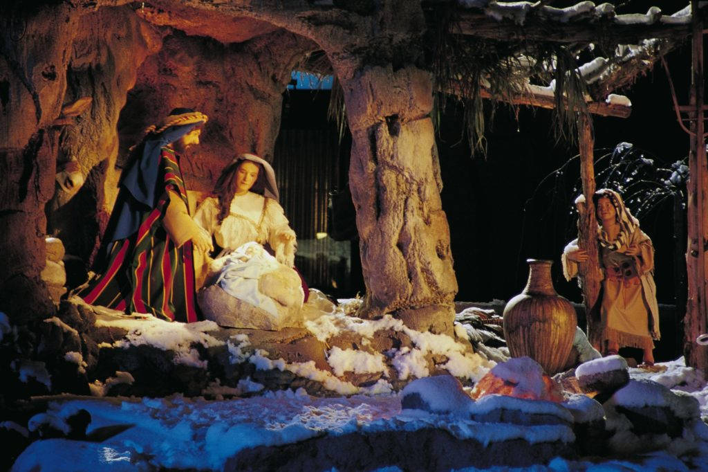 Nativity 桌布 壁紙 — 一個溫暖照明的耶穌誕生場景，瑪利亞、約瑟和嬰兒耶穌在一個被雪覆蓋的洞穴中，周圍是質樸的人物和陶器。