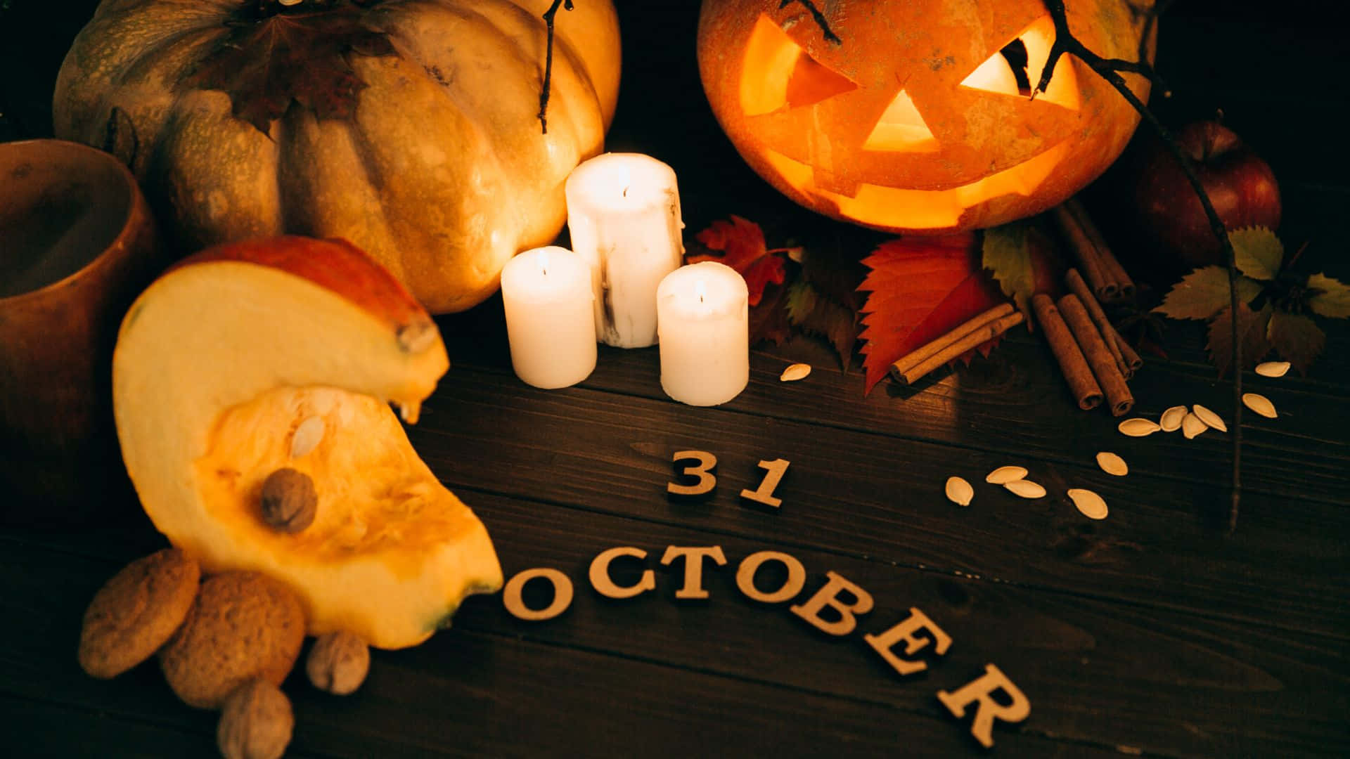 October 31st 桌布 壁紙 — 一個溫暖照明的萬聖節場景，有雕刻南瓜蠟燭、秋葉和深色木桌上的 '31 OCTOBER' 字母。